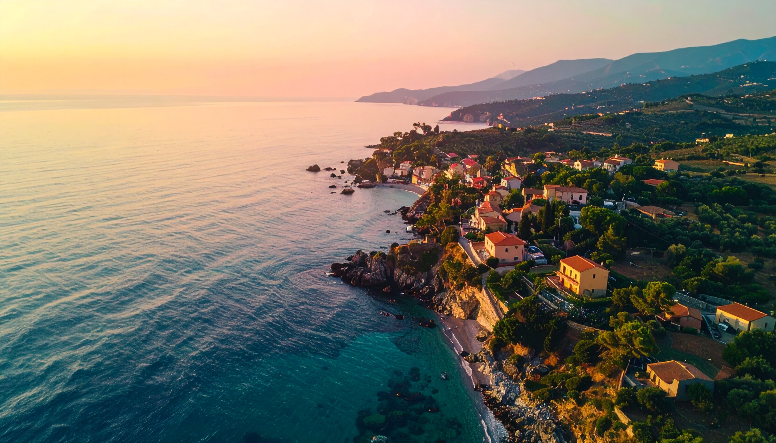 Vista aérea de uma pitoresca vila costeira ao entardecer, com casas coloridas distribuídas ao longo da costa rochosa. O mar calmo reflete tons alaranjados e rosados do pôr do sol, enquanto as colinas ao fundo adicionam profundidade à paisagem. A iluminação suave e natural acentua as texturas das árvores e das formações rochosas, criando uma atmosfera serena e bucólica.