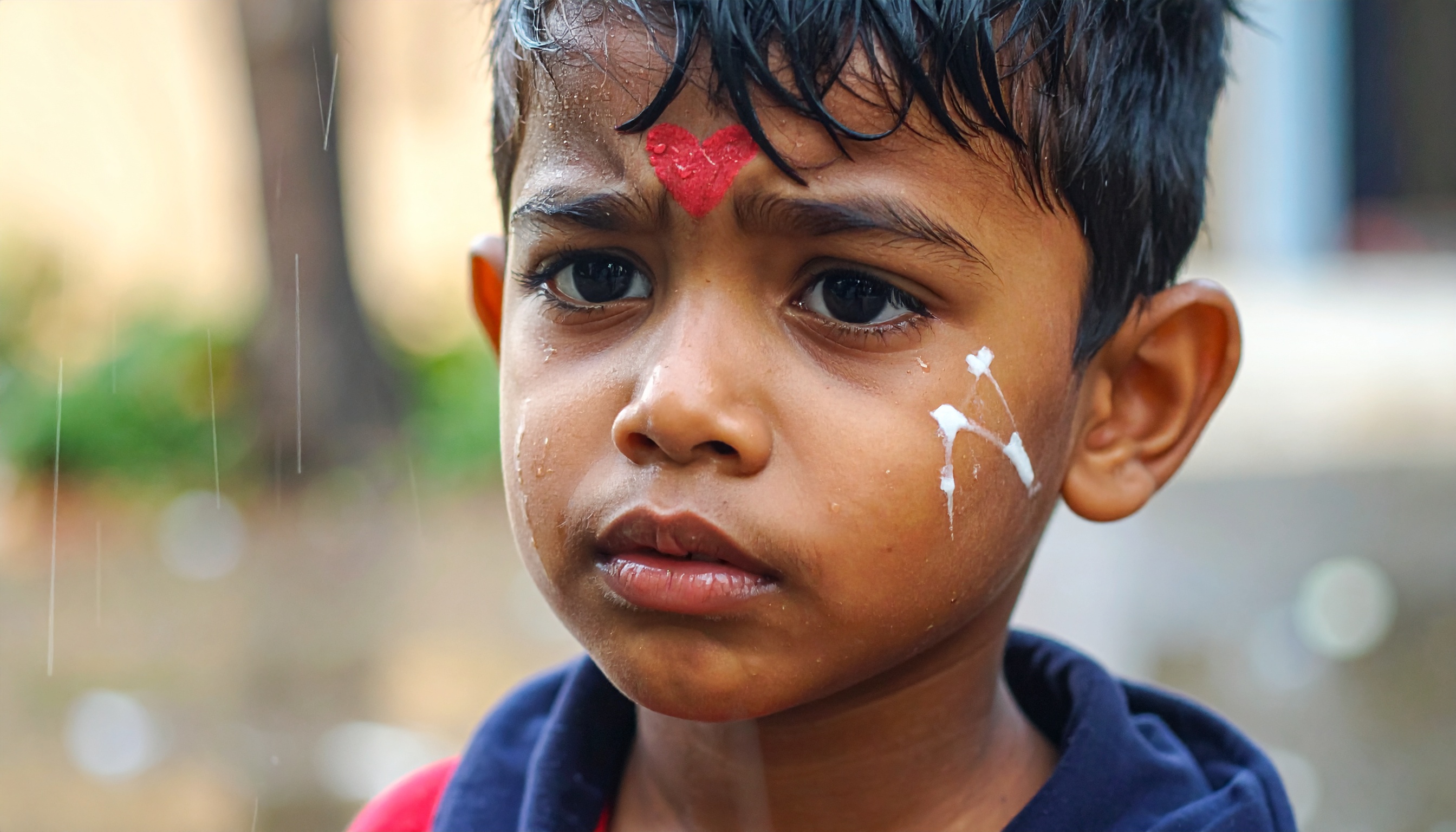 A young child with a painted forehead and cheeks stands outdoors, raindrops on their face