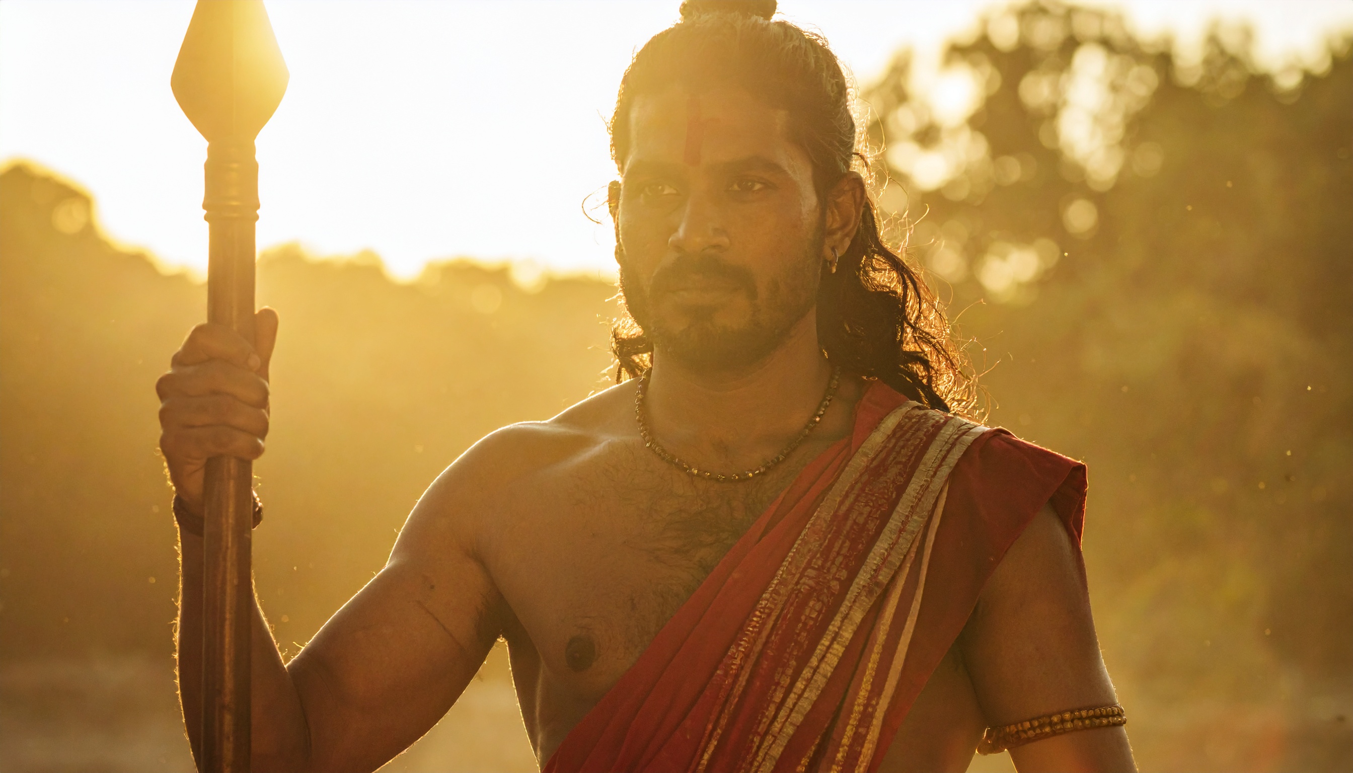 A man dressed in traditional attire holds a spear against a warm, golden sunset