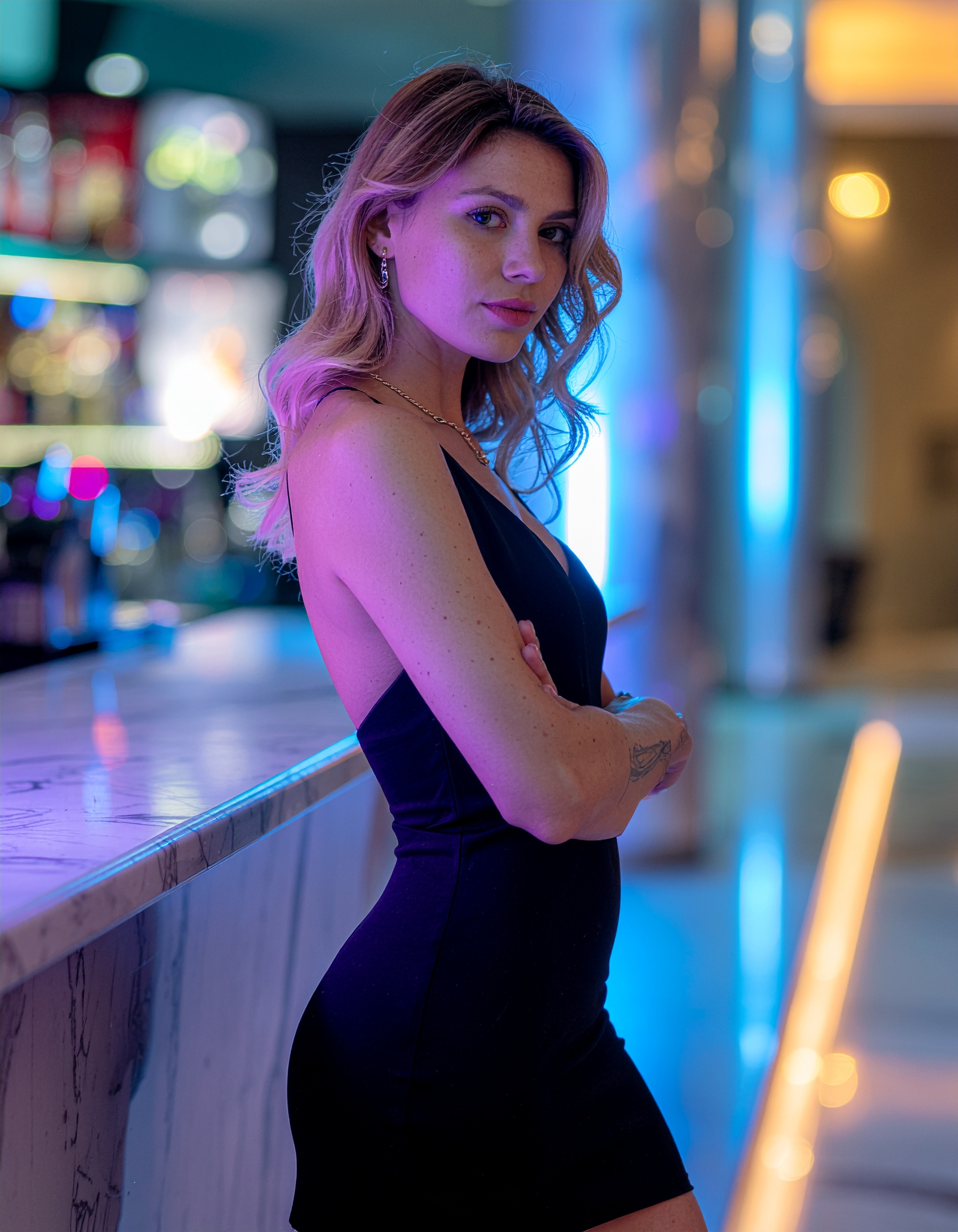 A woman in a stylish black dress stands by a modern marble bar with colorful lighting