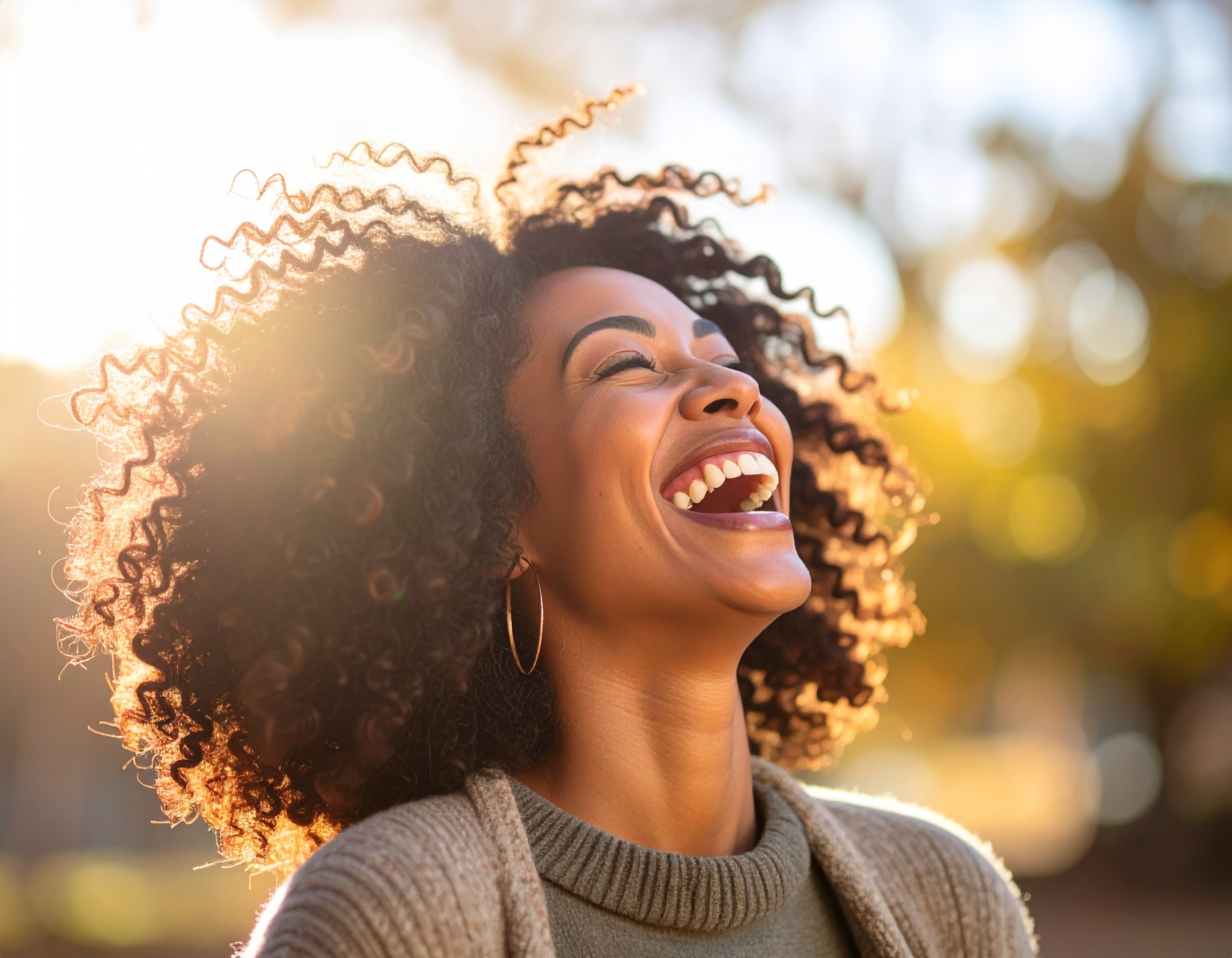 A woman with curly hair laughs joyfully in a sunlit outdoor setting