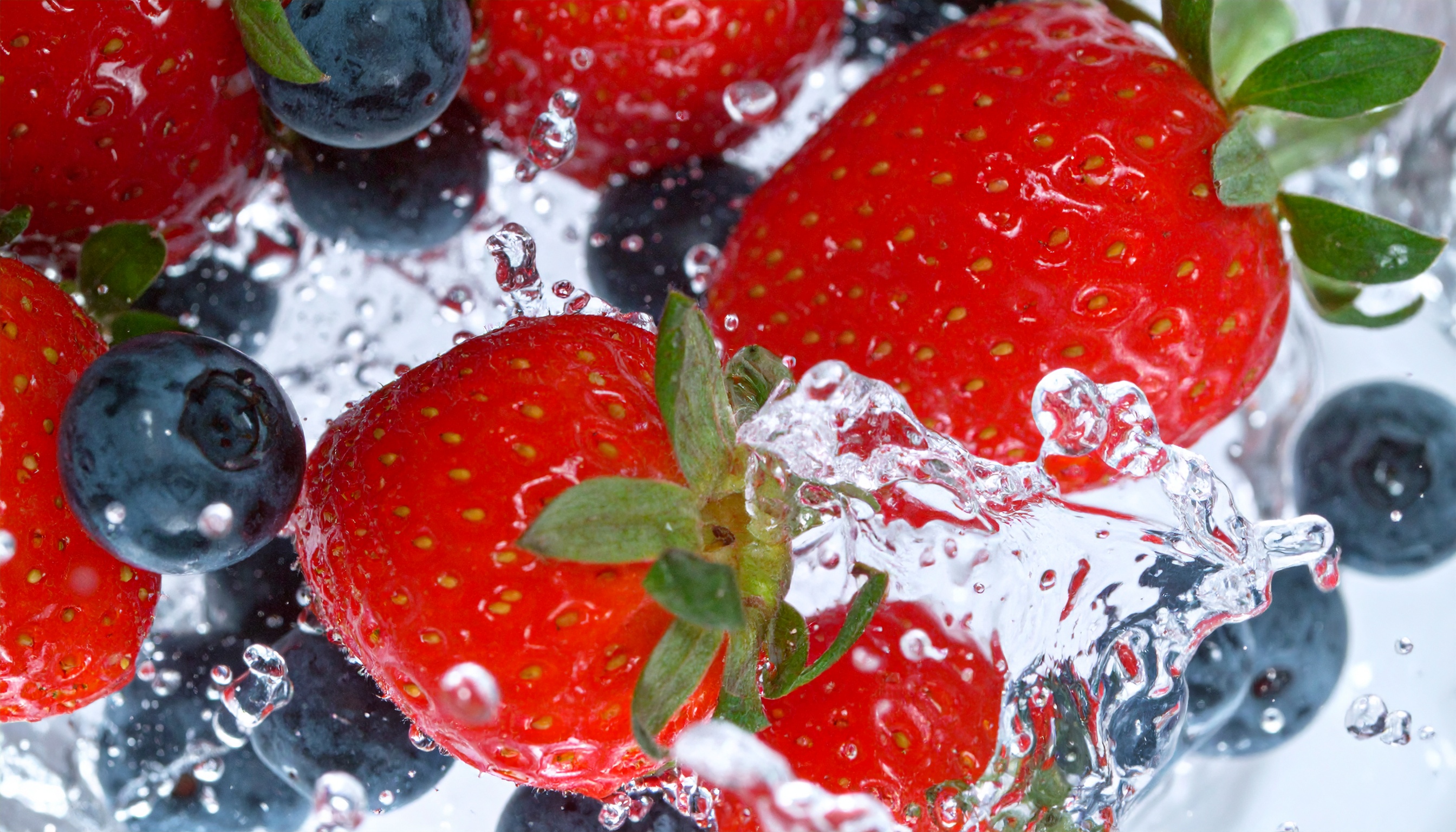 Imagem vibrante de morangos e mirtilos submersos em água cristalina, com gotas e respingos congelados no ar. As cores vivas das frutas contrastam com o fundo aquoso e translúcido. A composição é dinâmica, destacando a textura dos morangos e o brilho dos mirtilos, capturados em um close-up de alta definição que enfatiza a frescura e a vitalidade.