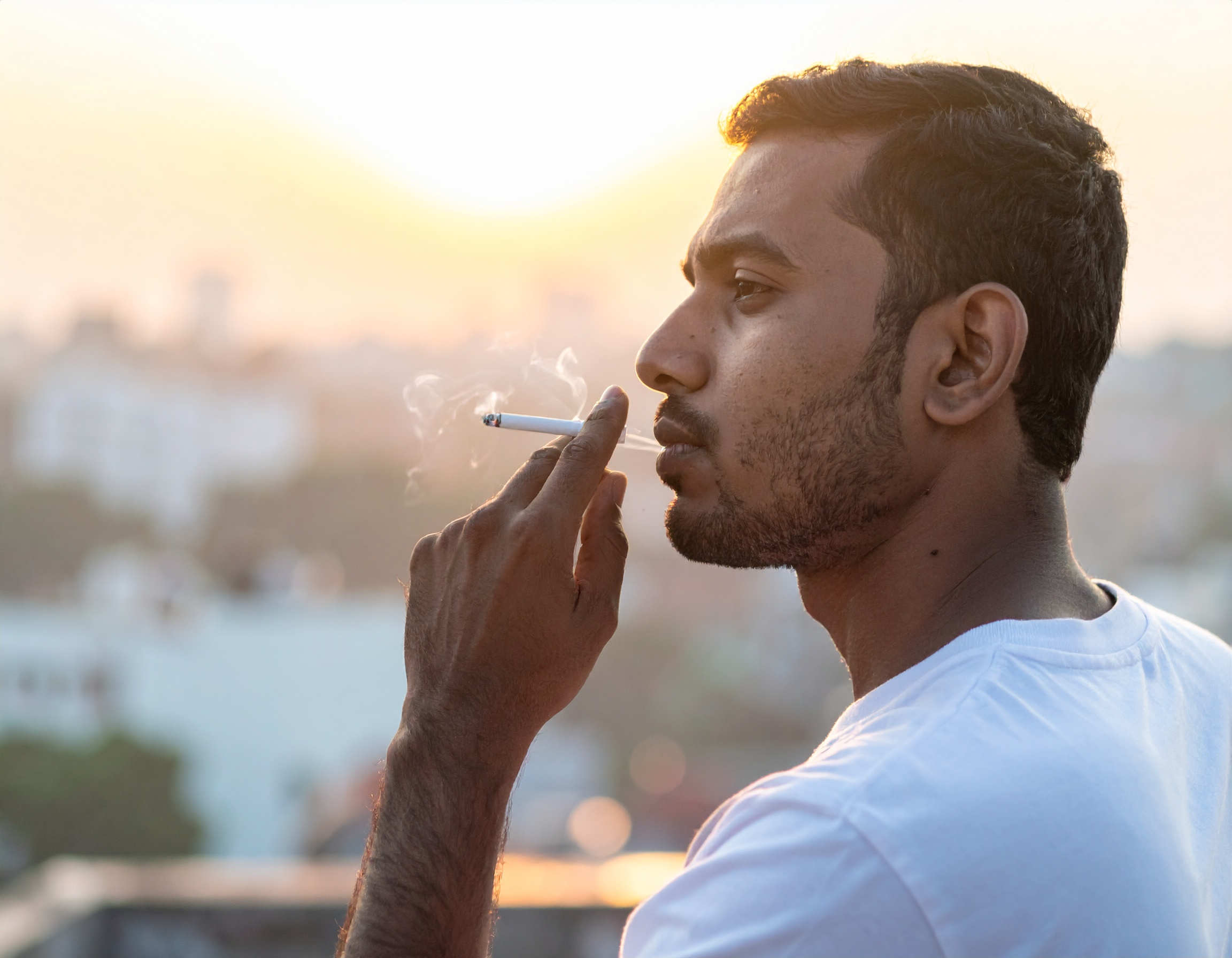 A man smokes a cigarette against a blurred urban backdrop