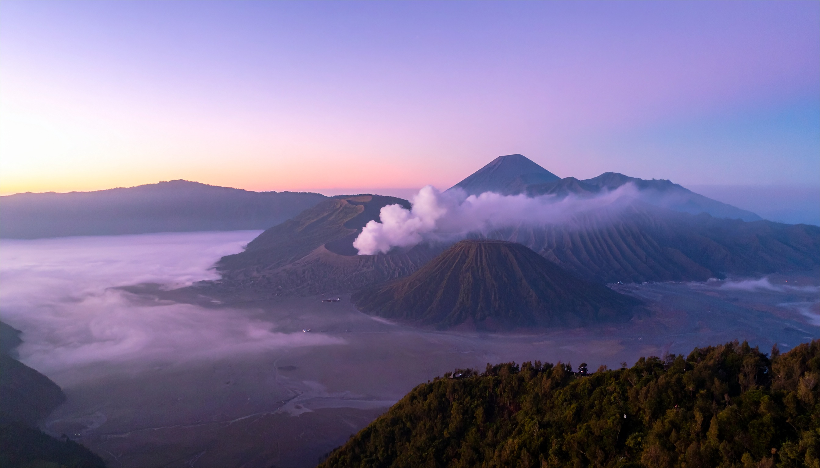 Amanhecer sobre o Monte Bromo, um vulcão icônico na Indonésia cercado por névoa. Localizado na ilha de Java, é popular entre turistas que buscam aventura e paisagens deslumbrantes. O vulcão exala uma fumaça suave, contrastando com as montanhas ao fundo e o céu colorido. Ideal para fotografias de viagem, expedições ao ar livre e inspiração na natureza. Esta paisagem vulcânica oferece uma visão única do poder da natureza e da serenidade matinal.