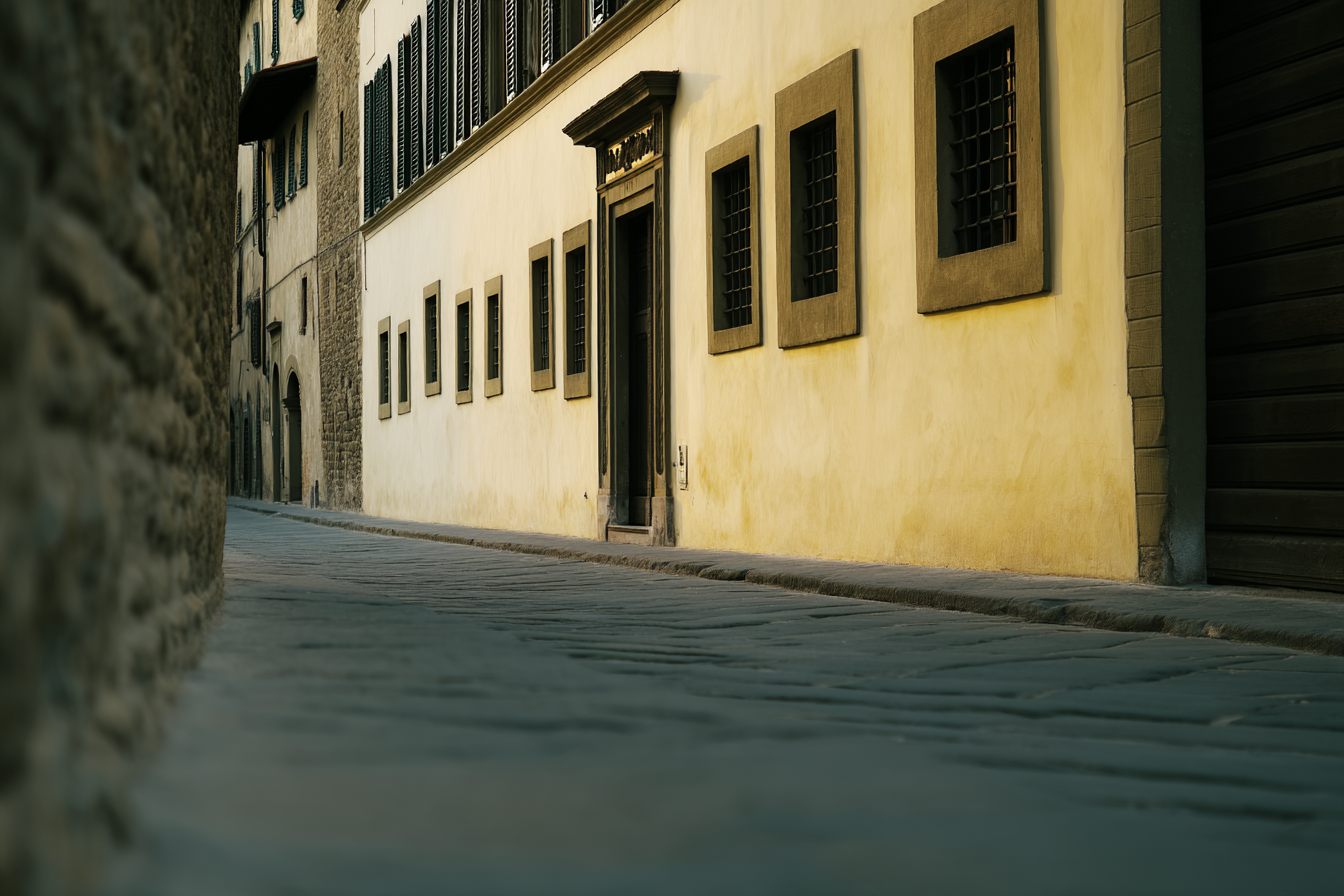 Rua estreita com fachadas de pedra e alvenaria amarelo-pálido, janelas com grades, portas de madeira escura, luz suave criando sombras sutis, atmosfera tranquila e histórica.