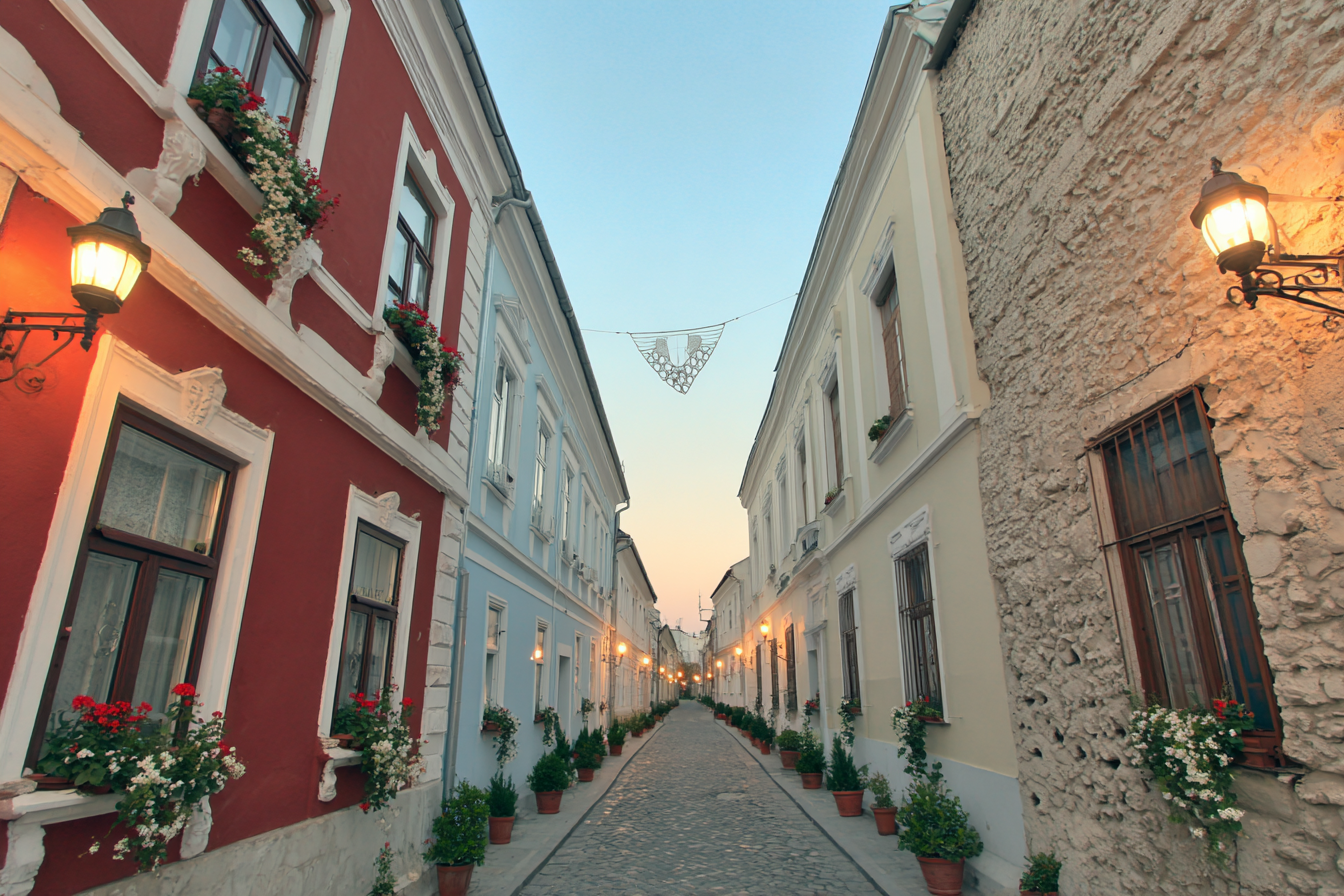 Rua estreita com calçamento de pedras, ladeada por edifícios históricos coloridos em tons de vermelho e azul claro. Janelas adornadas com flores pendentes e iluminação suave de postes antigos criam uma atmosfera encantadora ao entardecer, destacando a simetria e a profundidade da cena.