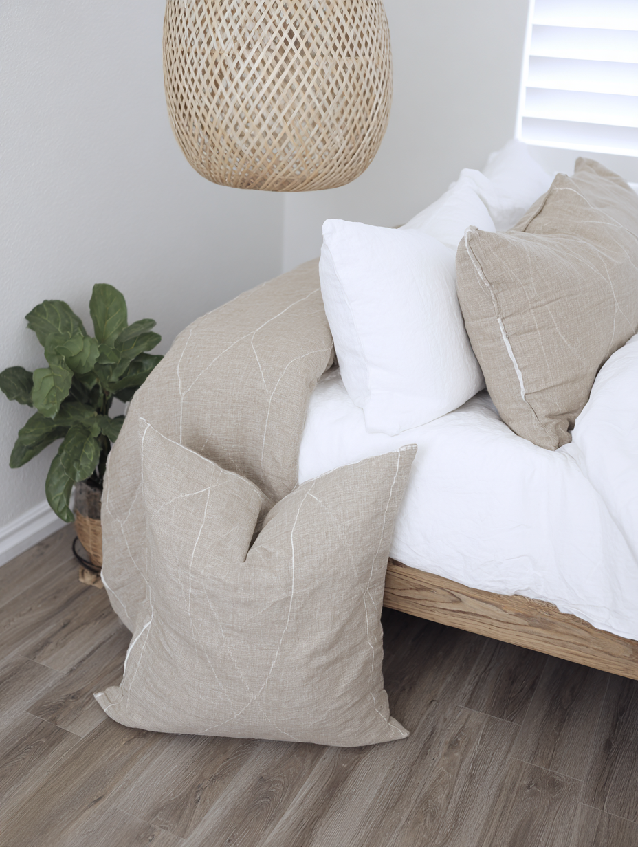 Quarto moderno com decoração minimalista, apresentando uma cama com lençóis brancos e almofadas bege de linho, ao lado de uma planta em vaso. O piso é de madeira clara, complementado por uma luminária de vime pendente, criando uma atmosfera serena e acolhedora. A luz natural suave entra pelas janelas, realçando a textura dos materiais e a harmonia das cores neutras.