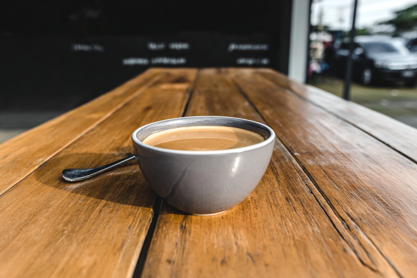 Uma xícara de café em cerâmica cinza descansa sobre uma mesa de madeira rústica iluminada pela luz natural, criando um ambiente acolhedor e sereno em um café ao ar livre.
