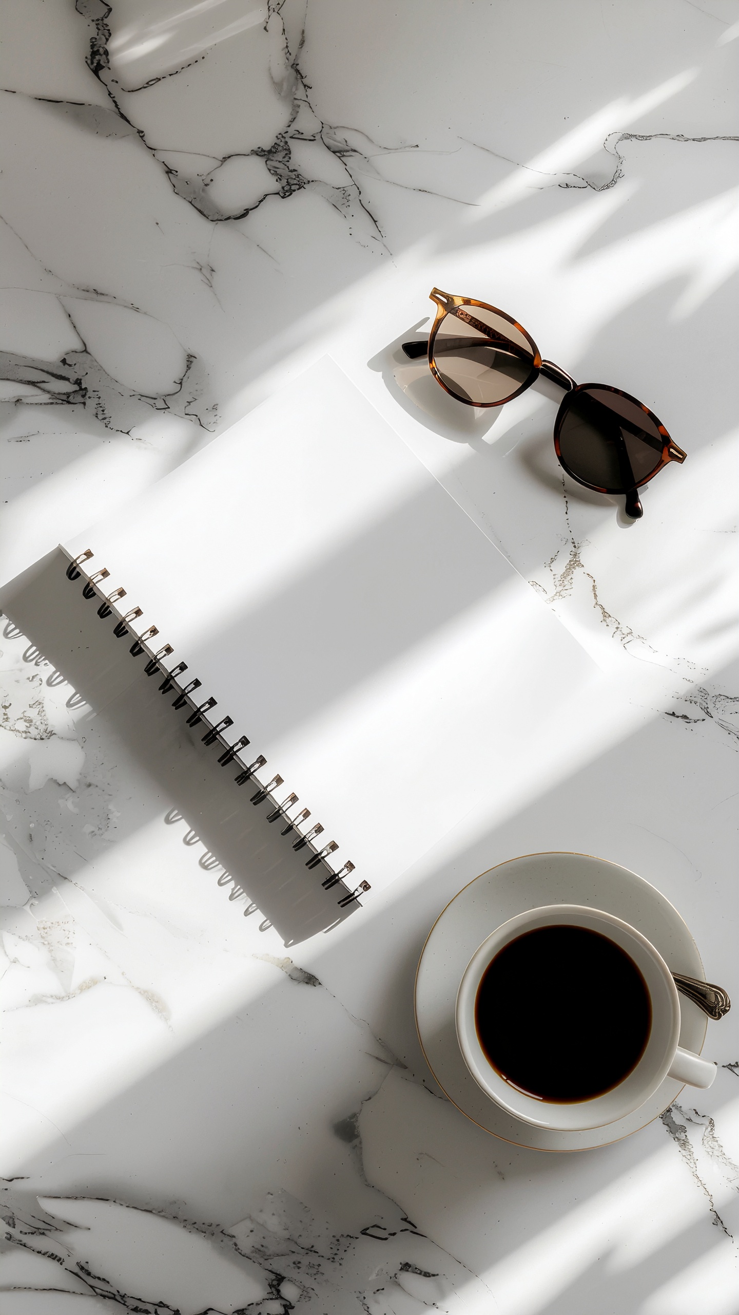 Cena minimalista com óculos de sol ao lado de uma xícara de café sobre uma mesa de mármore branco. Um caderno espiral está fechado, sugerindo um momento de pausa. A iluminação suave cria sombras delicadas, conferindo profundidade e textura à composição estética e elegante.