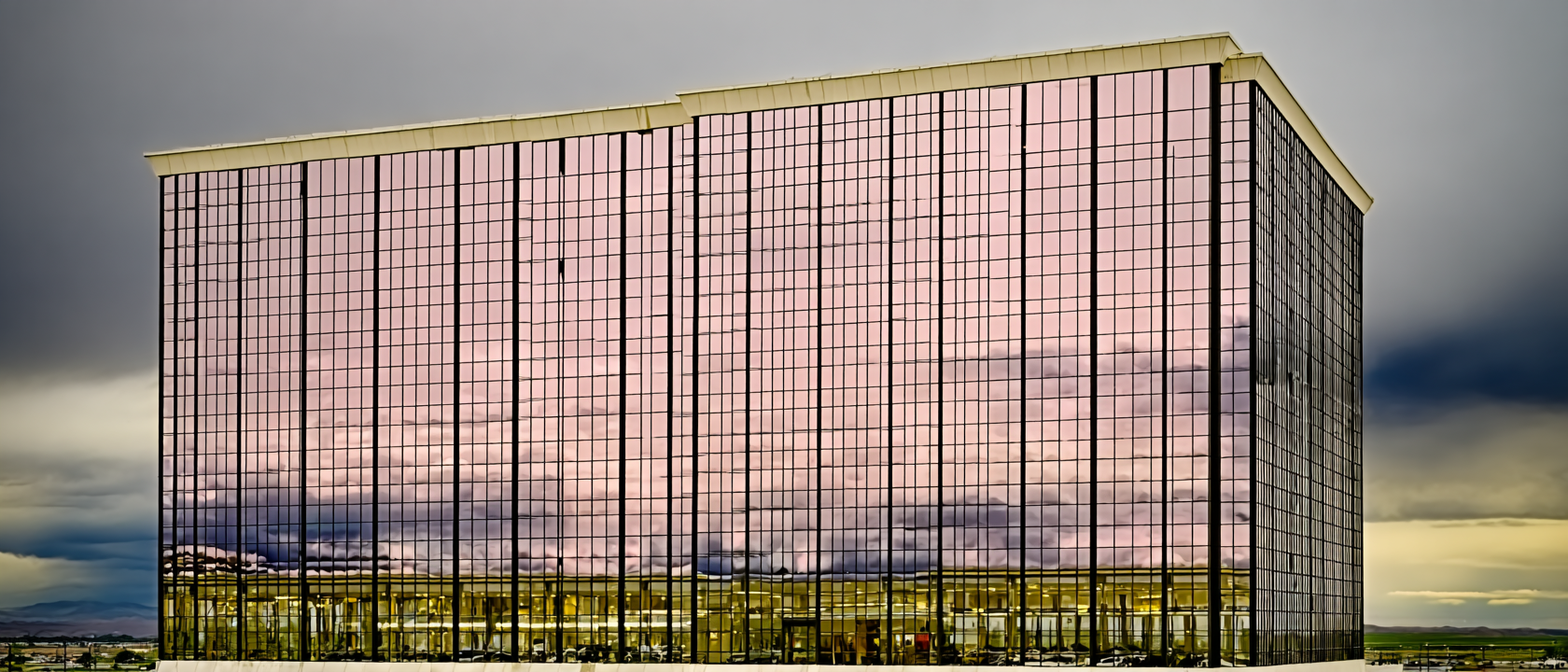 Edifício moderno com fachada de vidro refletindo o céu nublado, criando uma superfície espelhada que mistura tons de rosa e cinza. A estrutura quadrada e imponente possui linhas retas e simétricas, destacando-se contra o céu dramático. A iluminação suave e difusa do entardecer adiciona um toque de serenidade à cena urbana.