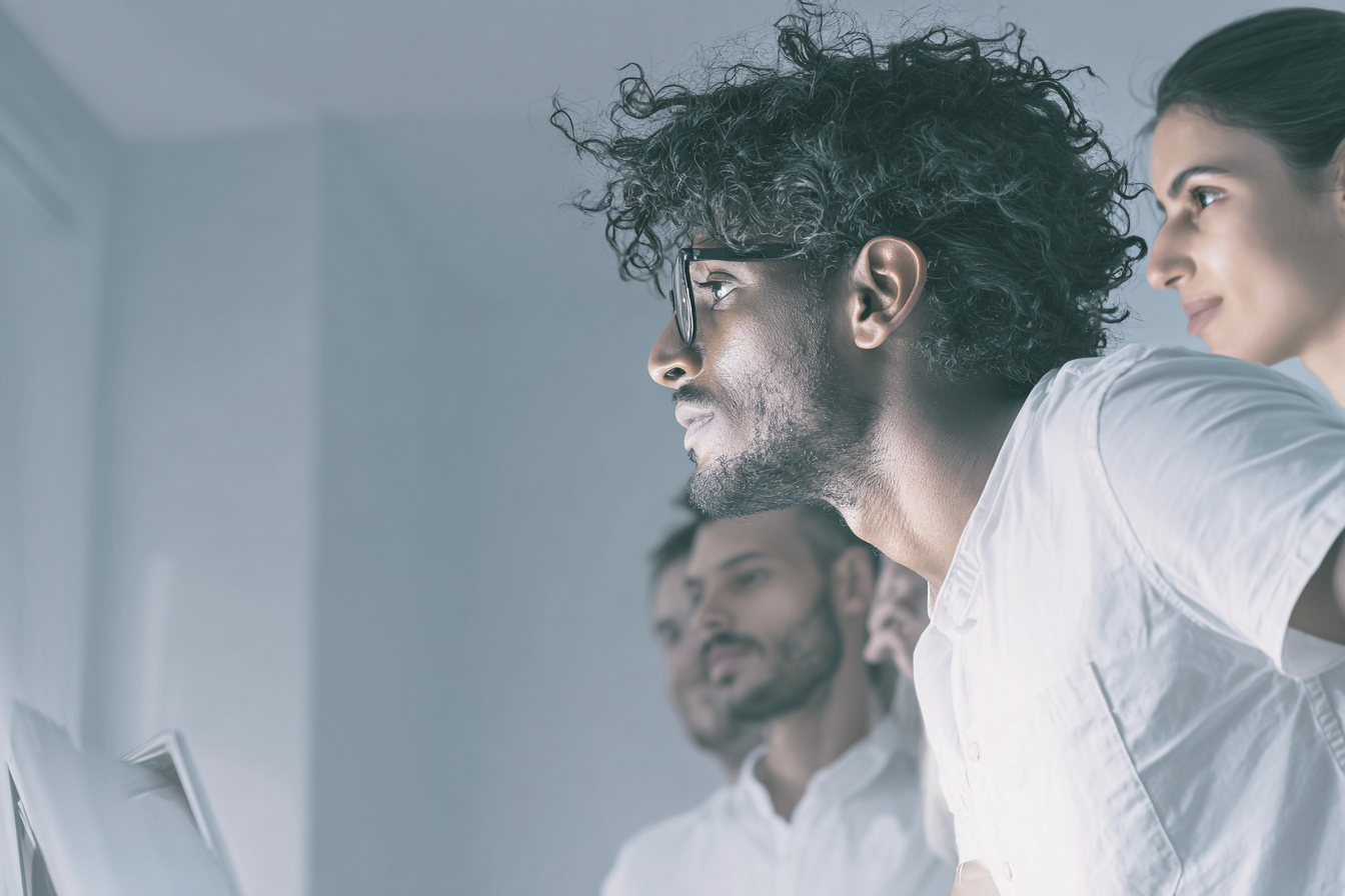 Três pessoas em um ambiente de trabalho moderno, focadas em uma tela fora de cena. A iluminação suave e fria destaca os detalhes faciais e a textura das roupas claras, transmitindo concentração e colaboração. A composição mostra foco e profundidade, com desfoque suave no fundo.
