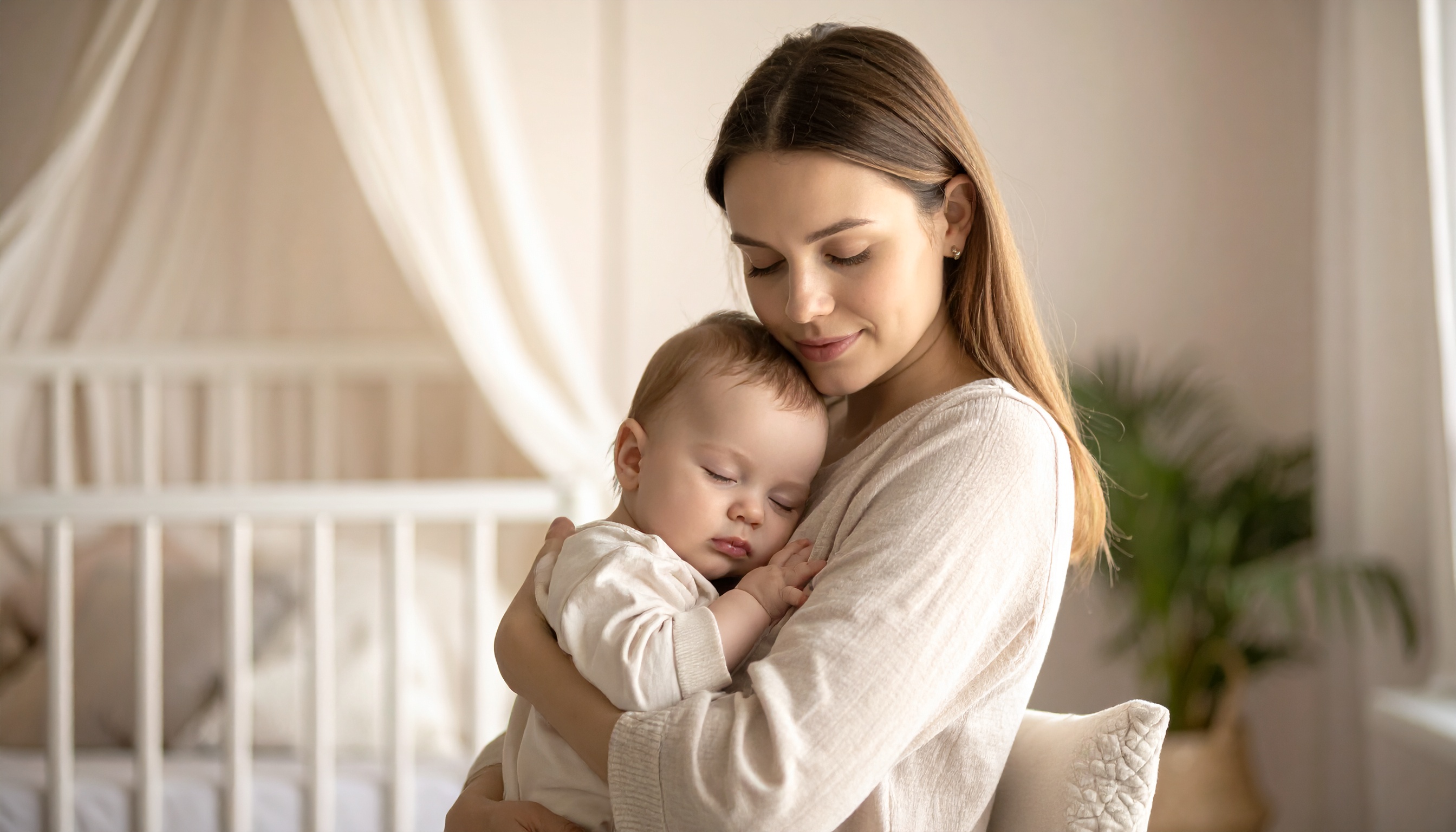 Uma mulher segura carinhosamente um bebê adormecido em um quarto iluminado e sereno, com tons suaves e uma atmosfera acolhedora. O berço ao fundo e a cortina branca criam um ambiente tranquilo. A imagem é capturada em um ângulo íntimo, destacando a expressão serena da mulher e a tranquilidade do bebê. A composição é equilibrada, com foco suave e luz natural suave, criando uma cena de ternura e paz.