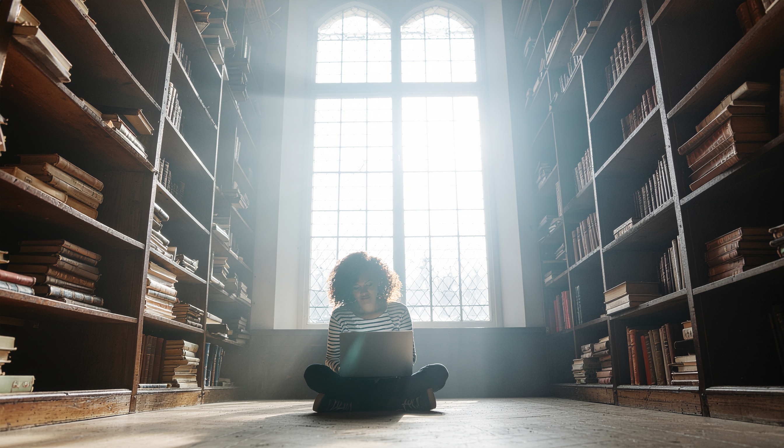Pessoa sentada no chão de uma biblioteca antiga iluminada por luz suave, cercada por estantes de livros, usando um laptop, transmitindo concentração e inspiração.