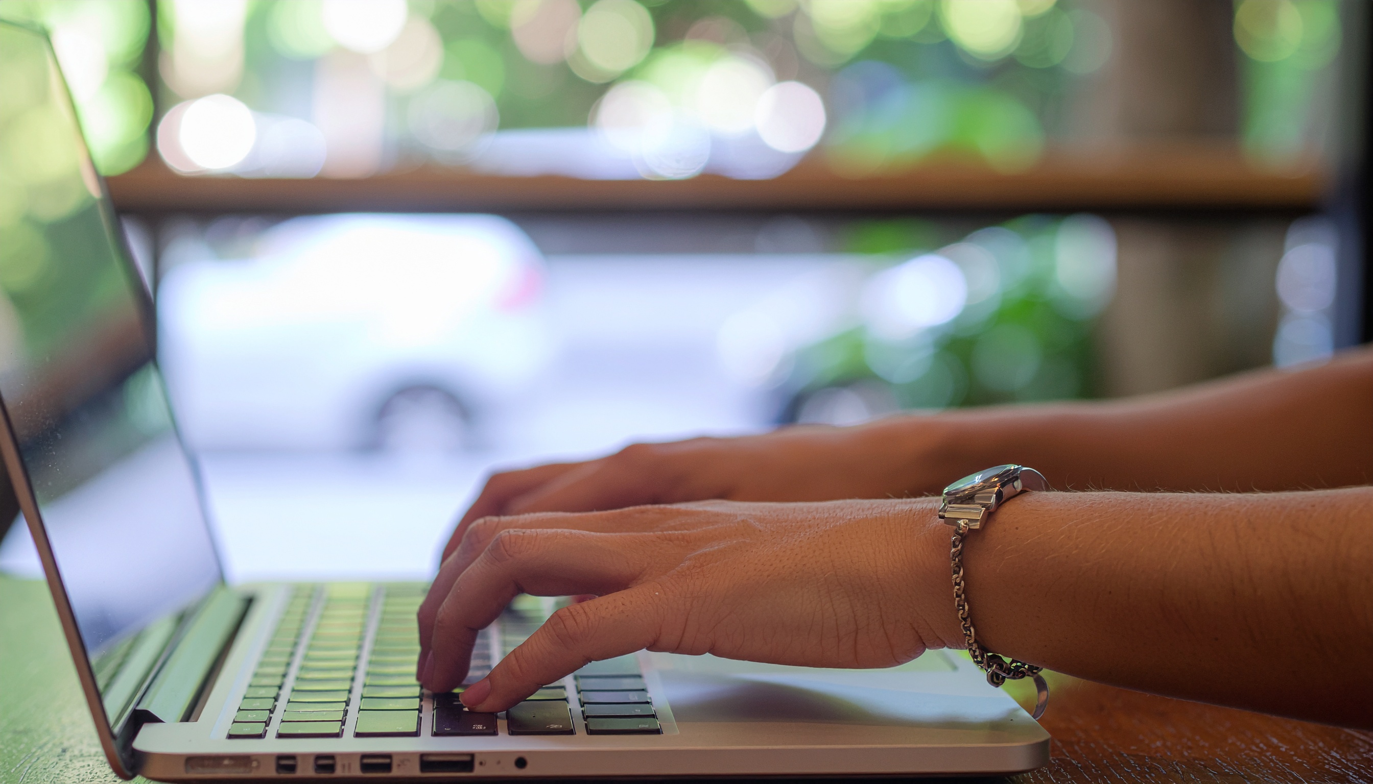 Mãos digitando em um laptop prateado em um ambiente iluminado por luz natural, com fundo desfocado de cores verdes e brancas, transmitindo uma atmosfera calma e produtiva. O foco está nítido nas mãos e teclado, enquanto o fundo permanece suavemente borrado, destacando a atividade principal.