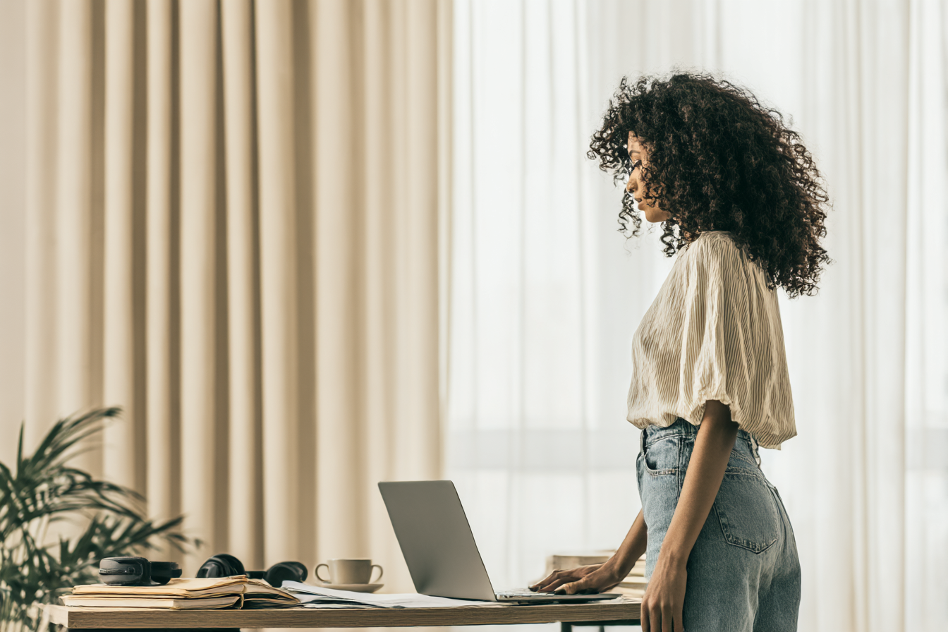Mulher de perfil com cabelo cacheado, usando uma blusa leve e jeans, trabalhando em um laptop sobre uma mesa de madeira em um ambiente iluminado por luz natural, com cortinas claras e plantas decorativas ao fundo.