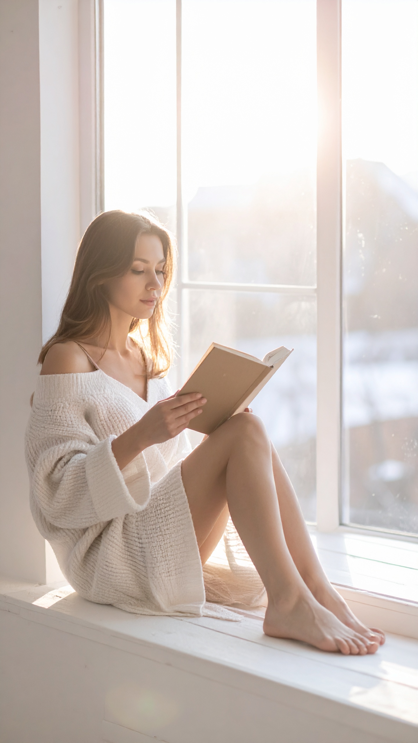 A woman in a cozy sweater sits by a sunlit window reading a book