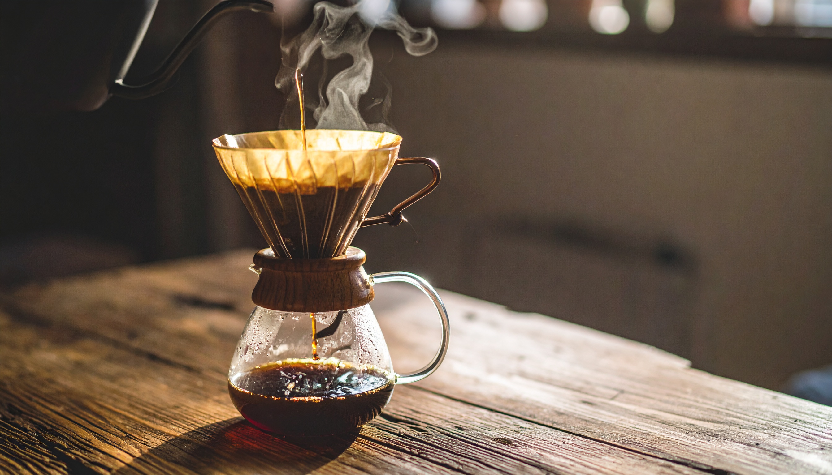 Café sendo preparado em um coador de vidro com filtro dourado, sobre mesa de madeira rústica. Iluminação suave destaca o vapor ascendente e o calor da cena, criando uma atmosfera acolhedora e artesanal.