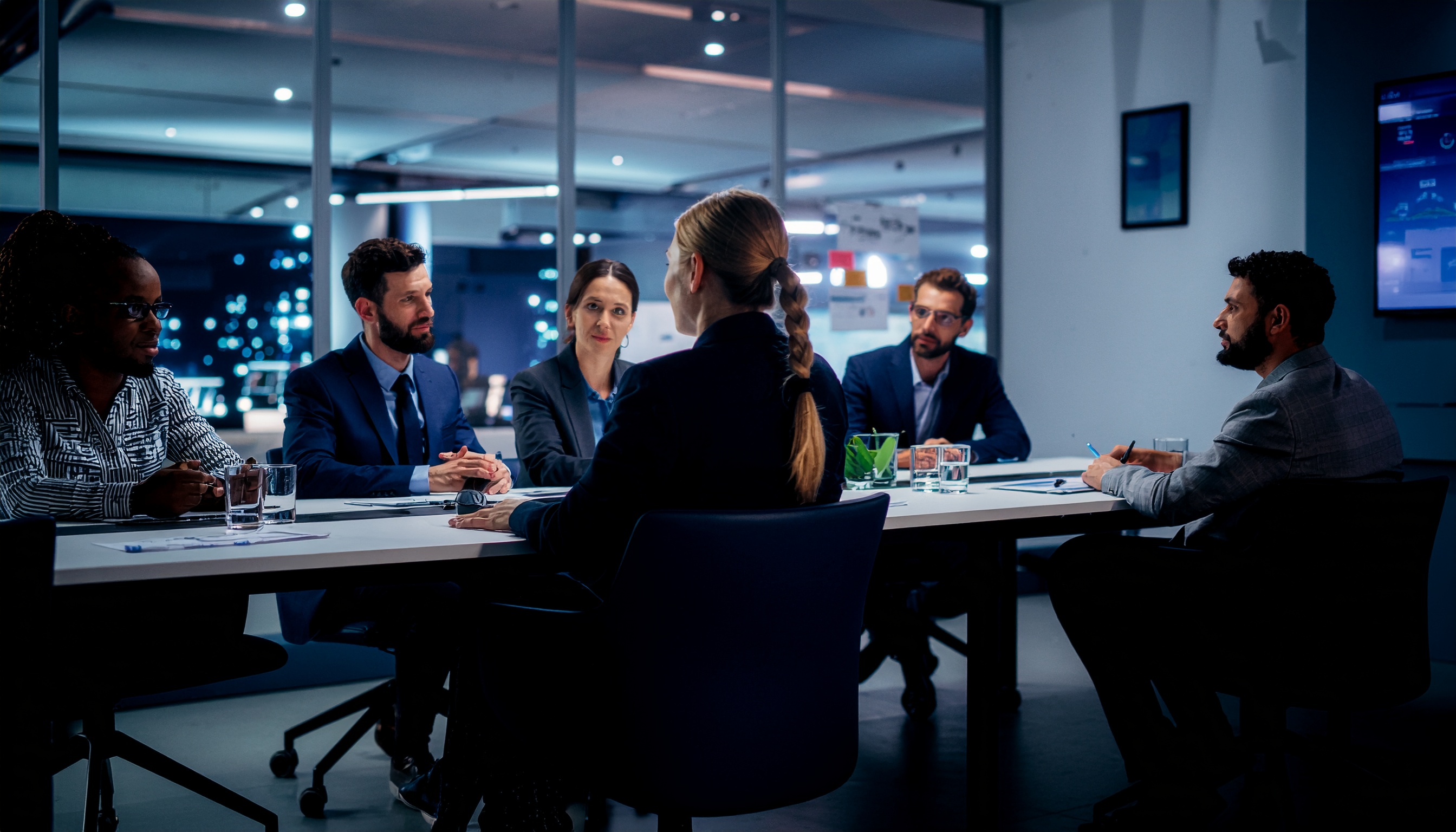 Reunião corporativa em uma moderna sala de conferências com seis profissionais discutindo estratégias sob iluminação suave. A atmosfera é de foco e colaboração.