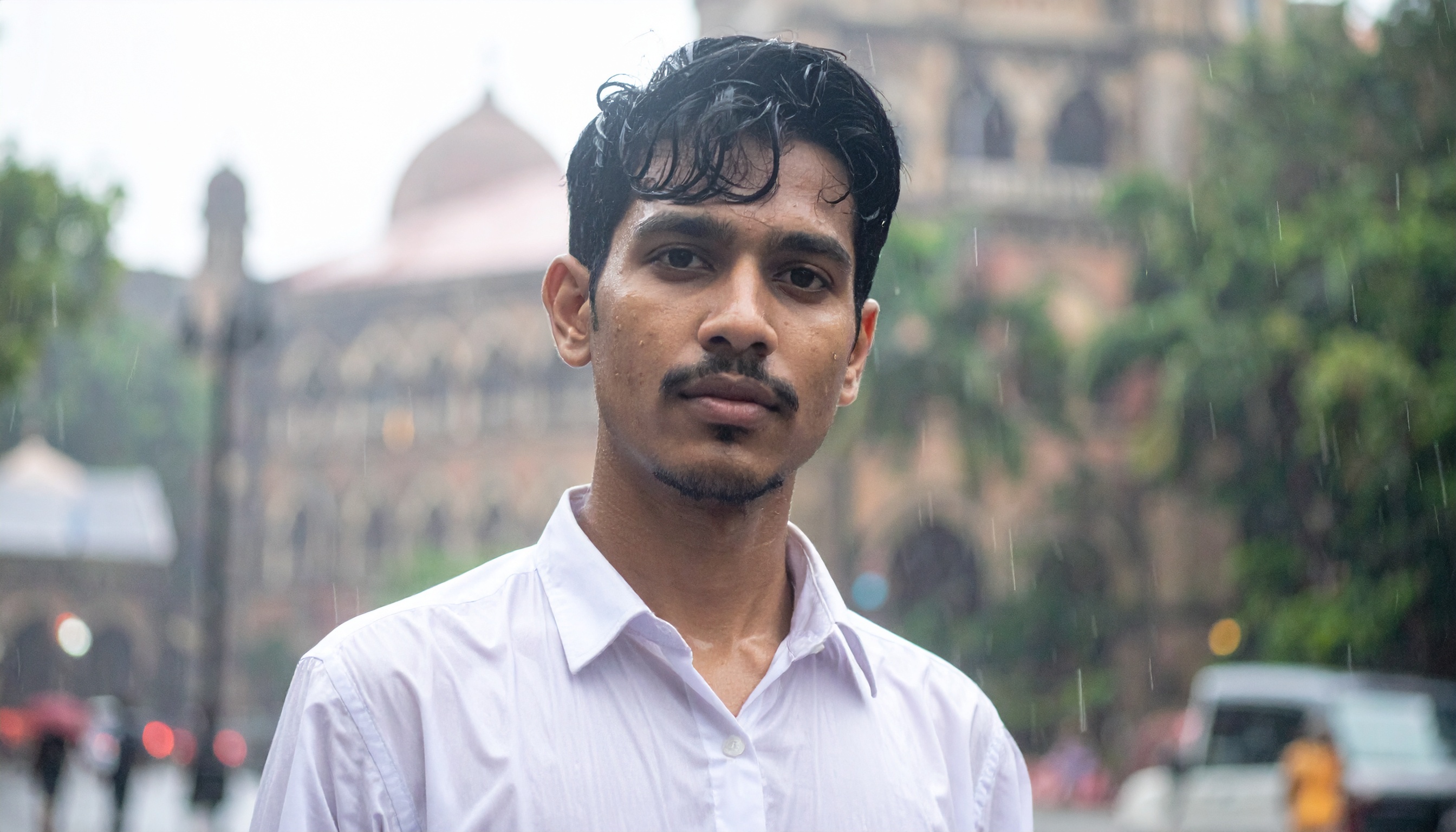 A man in a white shirt stands in front of a historic building on a rainy day