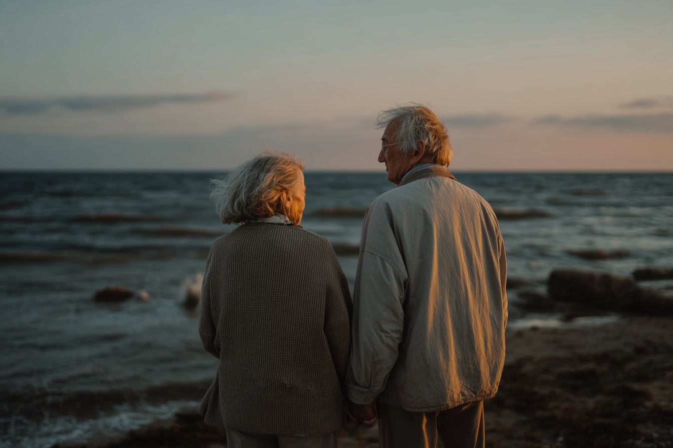 Um casal de idosos segurando as mãos à beira-mar durante o pôr do sol, com ondas suaves ao fundo e uma atmosfera tranquila e romântica.