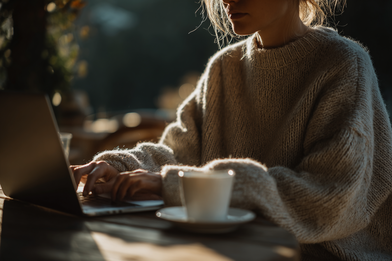 Mulher em suéter bege usando laptop em café aconchegante com luz suave da manhã, transmitindo tranquilidade e foco, ideal para trabalho remoto e estudo.