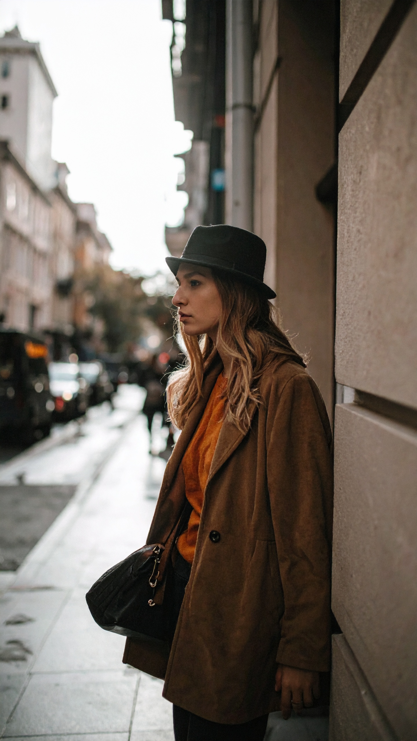 A stylish woman leans against a city wall wearing a chic brown coat and a black fedora