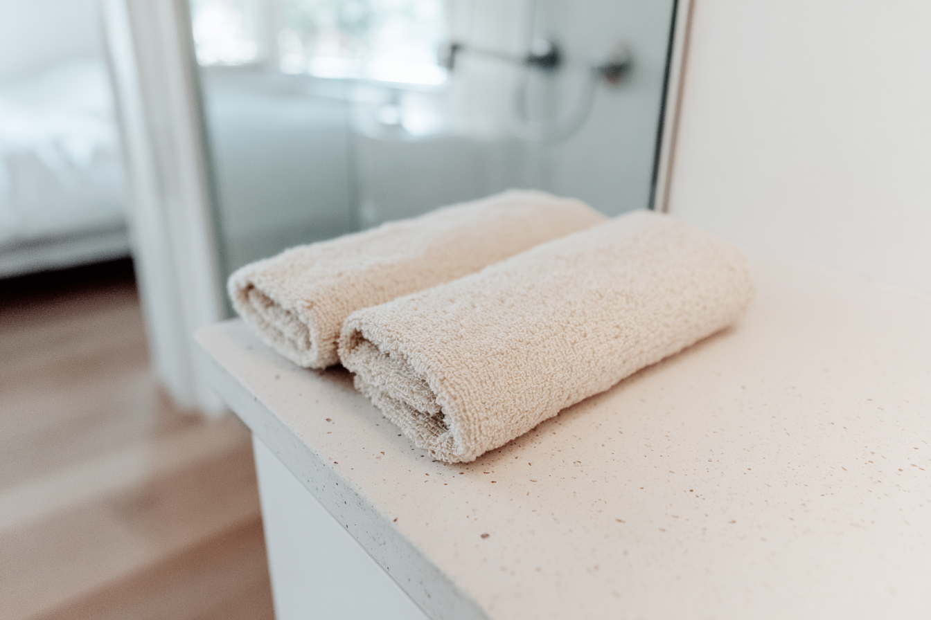 Dois toalhas de banho bege dobradas sobre uma bancada de banheiro clara, transmitindo sensação de limpeza e tranquilidade em um ambiente minimalista e bem iluminado.