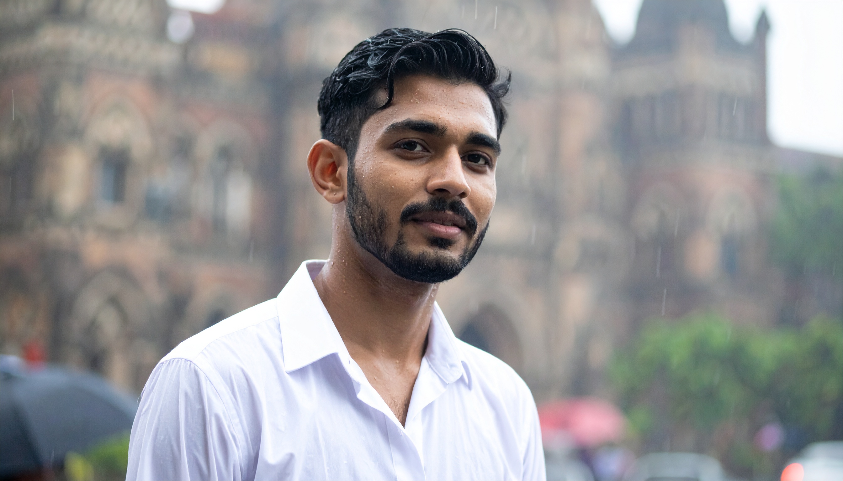 A young man stands confidently in front of a historic building, dressed in a crisp white shirt