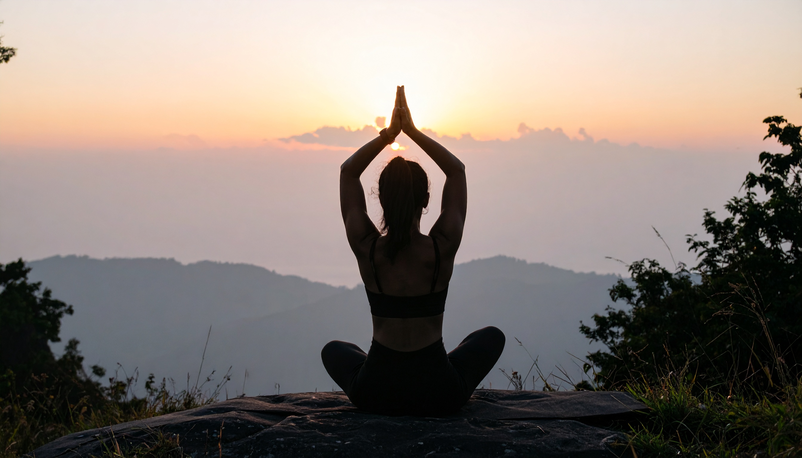 Silhueta de uma pessoa praticando yoga ao amanhecer em um cenário natural, com as montanhas ao fundo e um céu alaranjado suave. A postura meditativa é destacada pela iluminação suave e composição equilibrada, criando uma atmosfera de tranquilidade e introspecção. A imagem é capturada em ângulo reto, com foco na figura central contra o horizonte nebuloso.