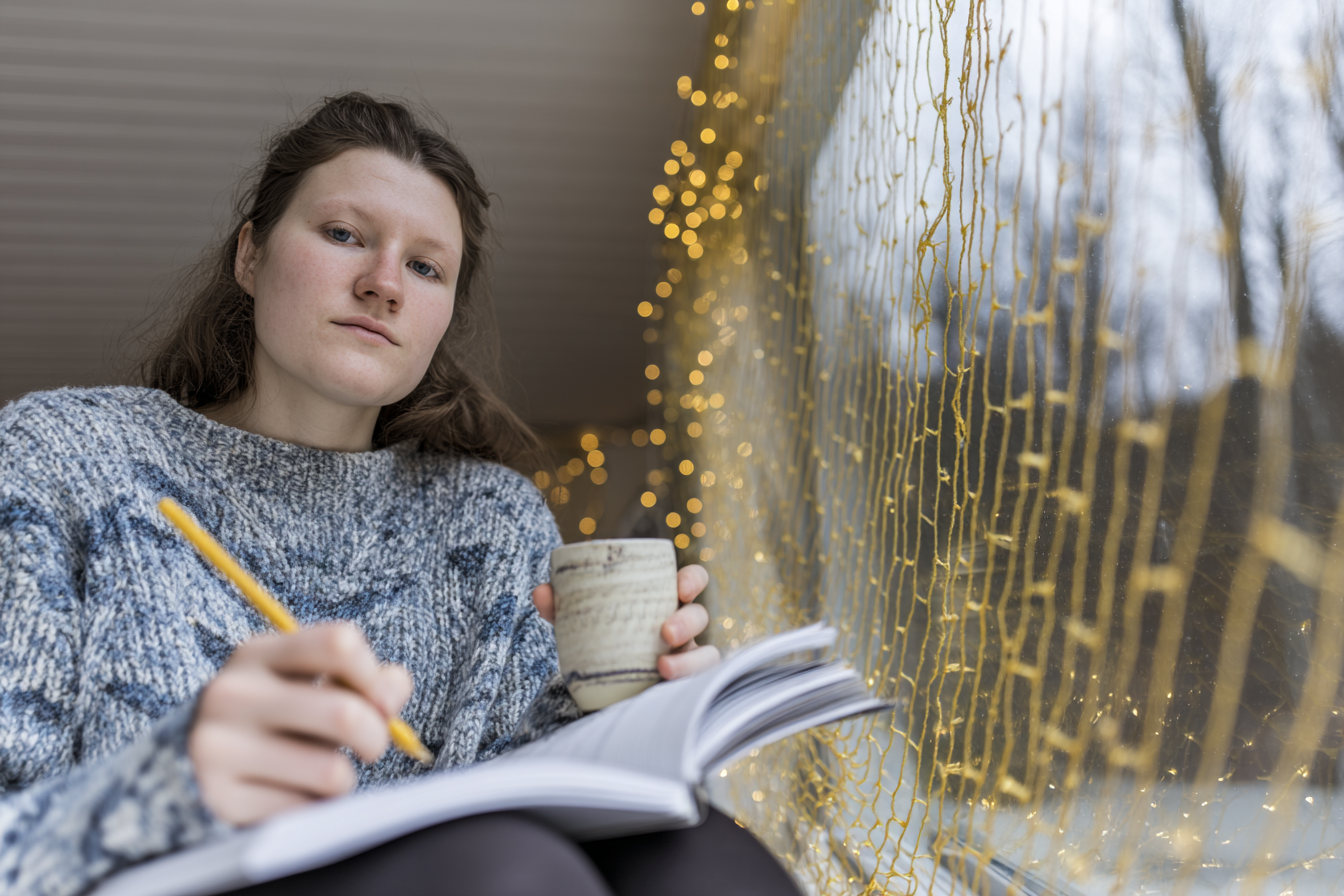 Jovem sentada ao lado de uma janela decorada com luzes amarelas, vestindo suéter cinza, segurando caneca e escrevendo em caderno. Cena iluminada suavemente com foco no rosto e detalhes da decoração.