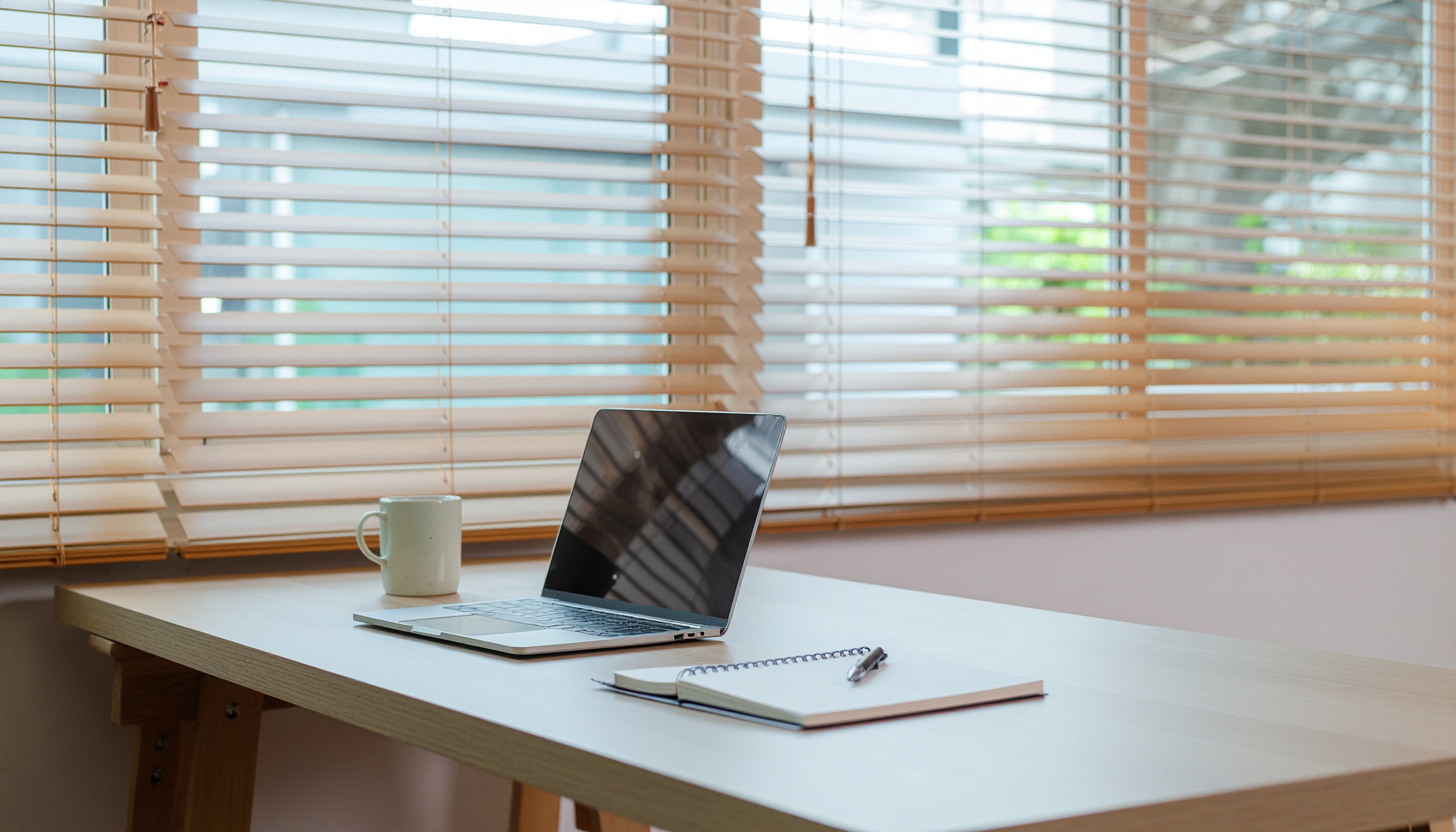 Mesa de escritório minimalista com laptop prateado aberto, caneca de cerâmica branca e caderno espiral, iluminada por luz suave através de persianas de madeira clara, criando uma atmosfera calma e organizada.
