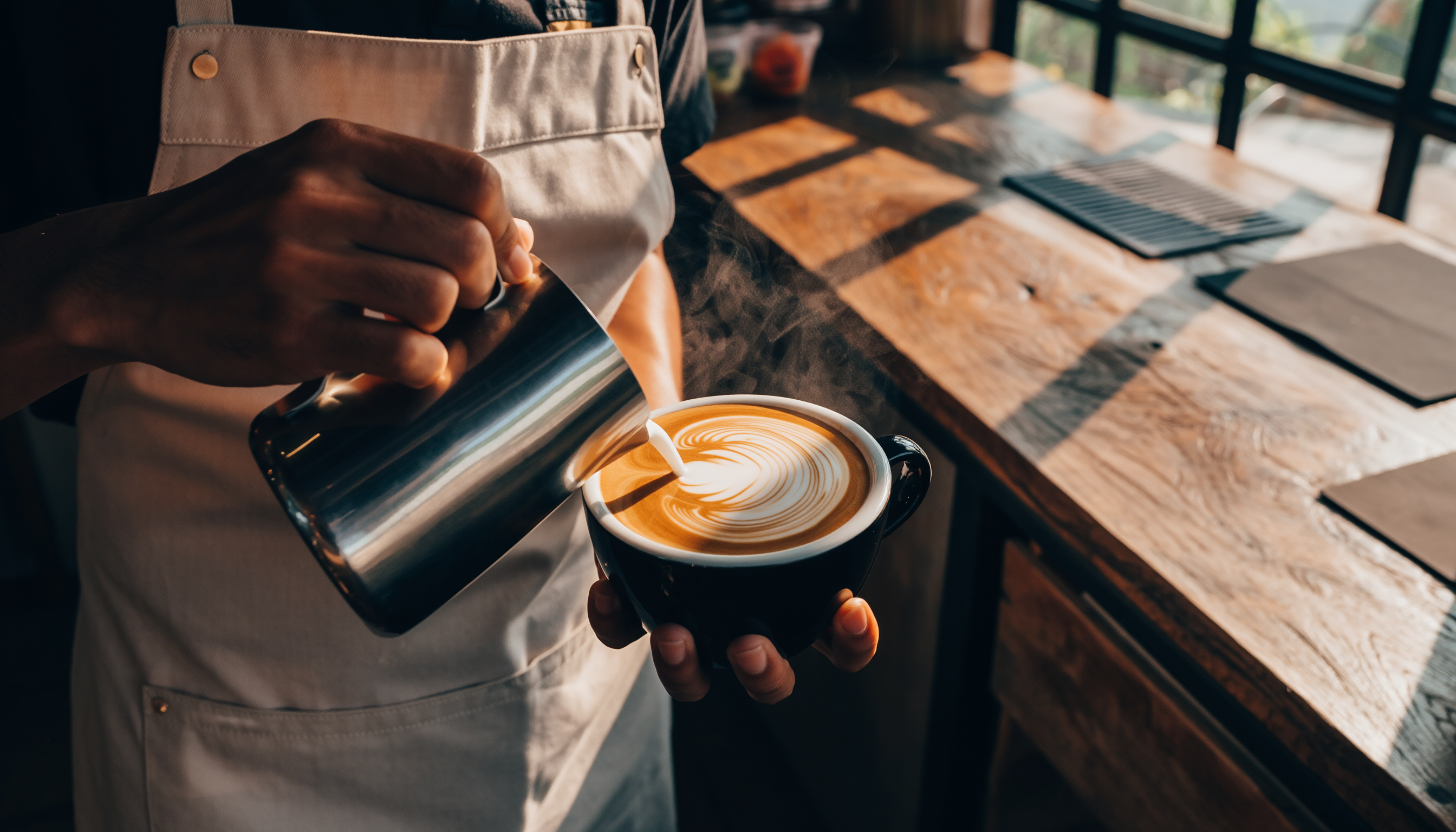 Barista vestindo avental branco derrama leite em um cappuccino, criando uma arte de latte. A iluminação suave destaca o vapor e o detalhe do desenho, em um ambiente acolhedor de cafeteria com luz natural entrando pela janela.