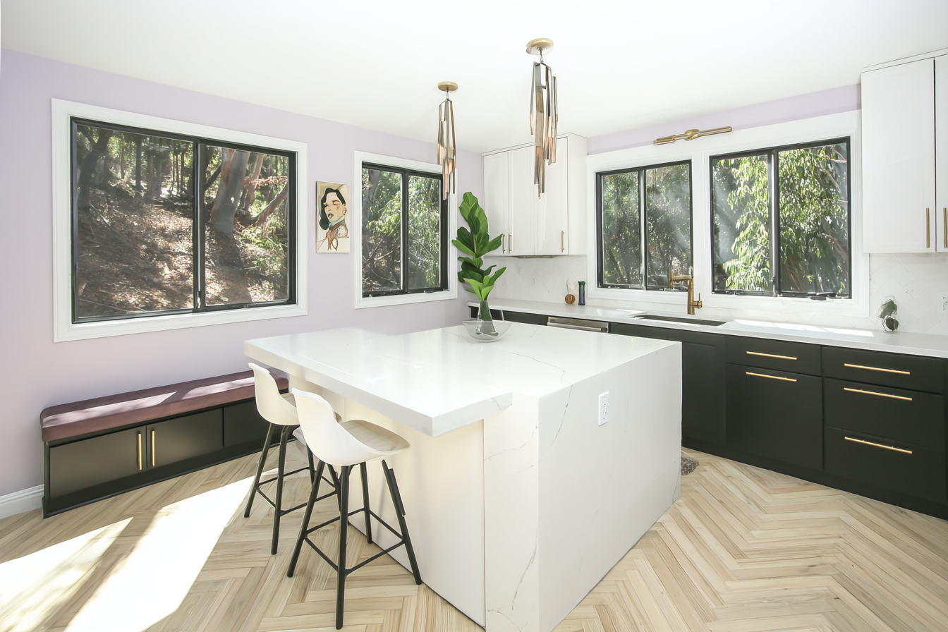 Cozinha moderna com iluminação natural, paleta clara com paredes lilás e bancada branca, piso em padrão espinha de peixe, decoração minimalista e janelas amplas.