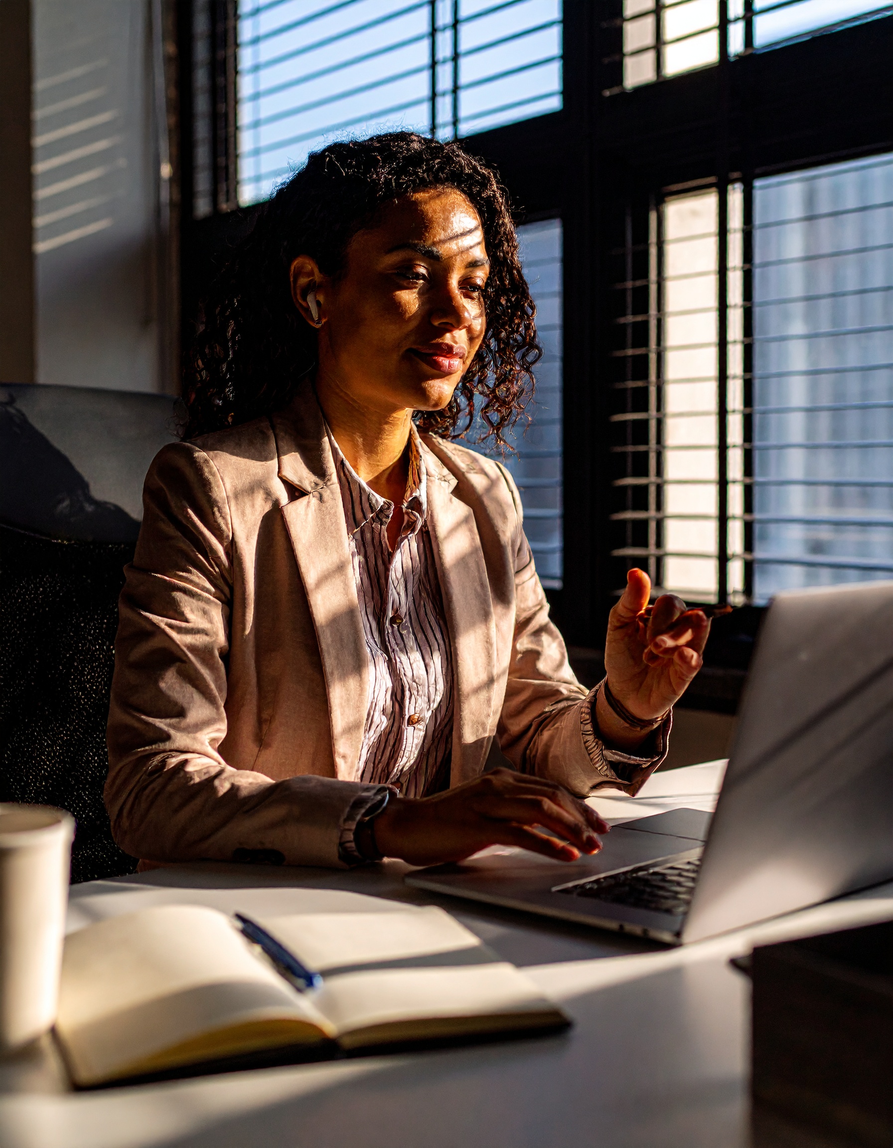 Mulher em escritório iluminado pelo sol poente, vestindo blazer bege com listras suaves, usando laptop. A luz cria padrões de sombra nas persianas.