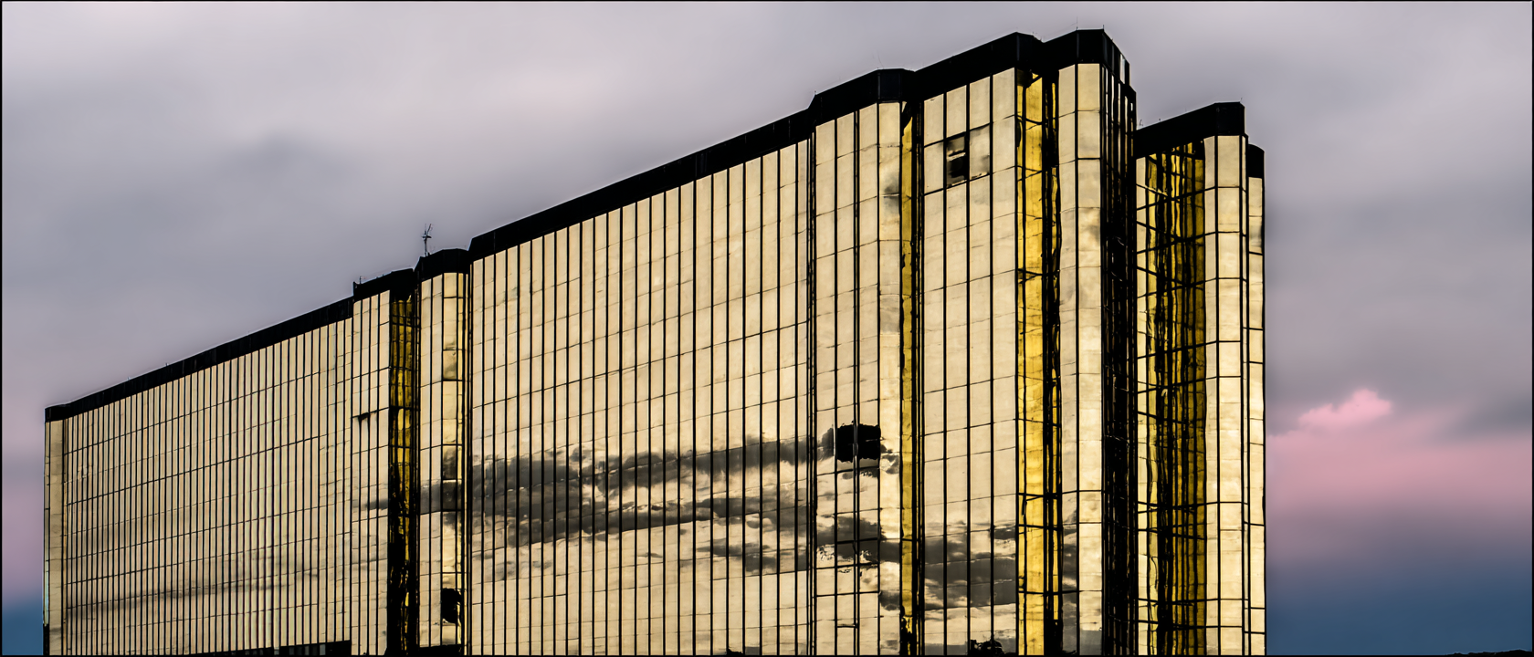 Edifício moderno com fachada espelhada dourada refletindo o céu nublado, destacando-se contra um fundo de cores suaves ao entardecer. As linhas retas e simétricas da arquitetura criam um efeito visual de profundidade, enquanto a luz dourada confere um toque de luxo e sofisticação. A composição é elegante e minimalista, capturada em ângulo que enfatiza a altura e imponência da estrutura.