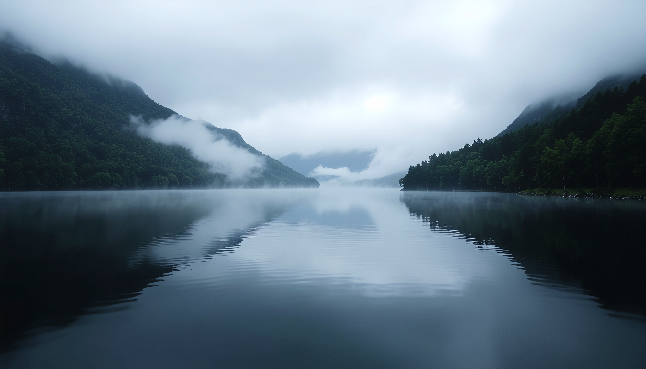 Paisagem serena com lago espelhado refletindo montanhas cobertas de vegetação e nuvens baixas, criando uma atmosfera de tranquilidade e mistério. A composição simétrica e a iluminação suave destacam a profundidade e a calma do cenário natural.