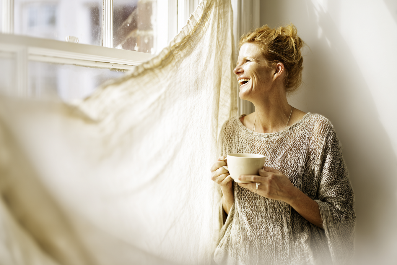 Mulher sorrindo alegremente enquanto desfruta de uma xícara de café, iluminada pela luz suave do sol através da janela. A atmosfera é acolhedora e tranquila, capturando um momento de paz e felicidade em um ambiente doméstico sereno.