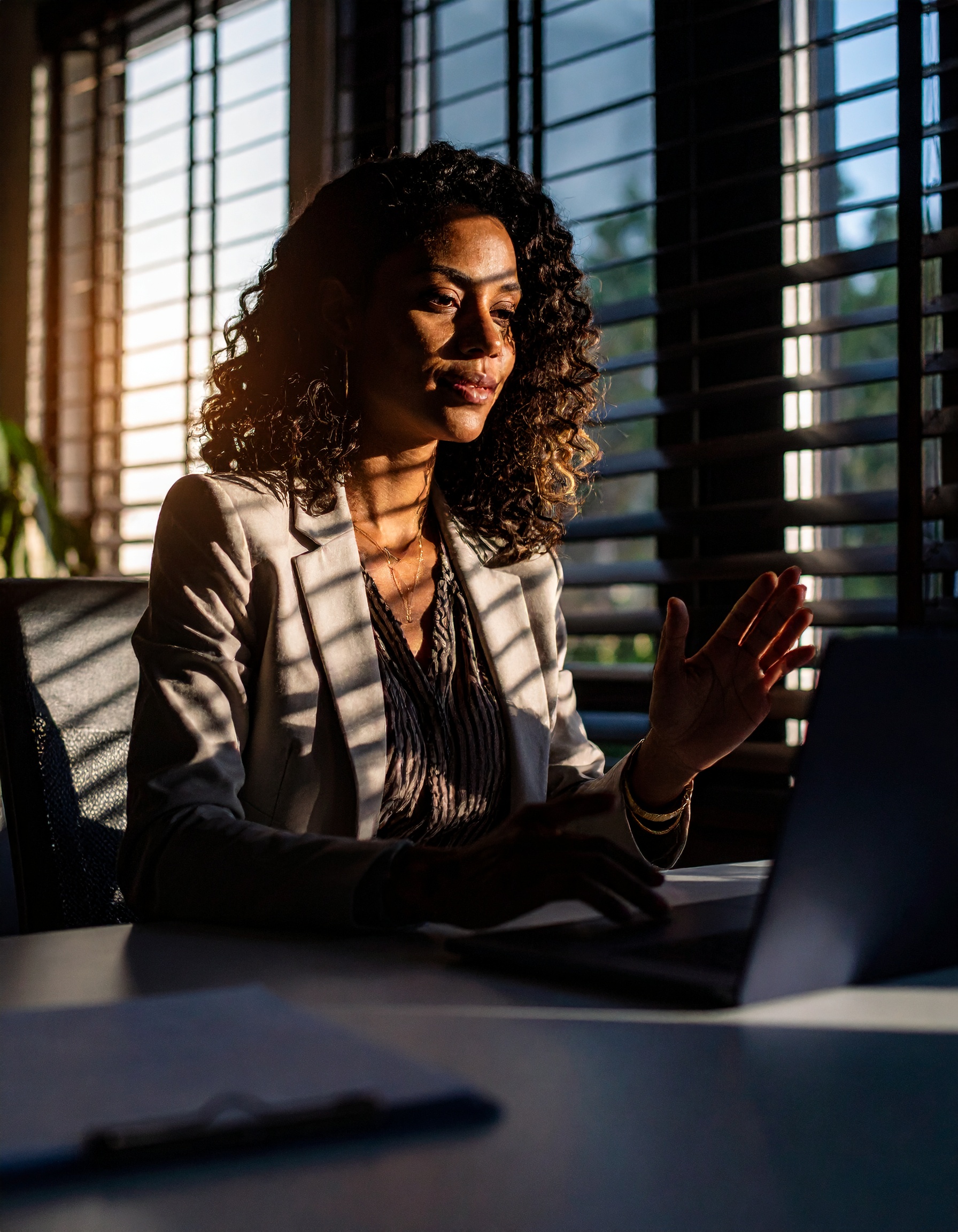 Mulher em ambiente de escritório, sentada à mesa com laptop, luz do sol filtrando pelas persianas, criando sombras dramáticas no rosto. A iluminação natural destaca o cabelo cacheado e a expressão focada, enquanto o fundo desfocado dá profundidade à cena.