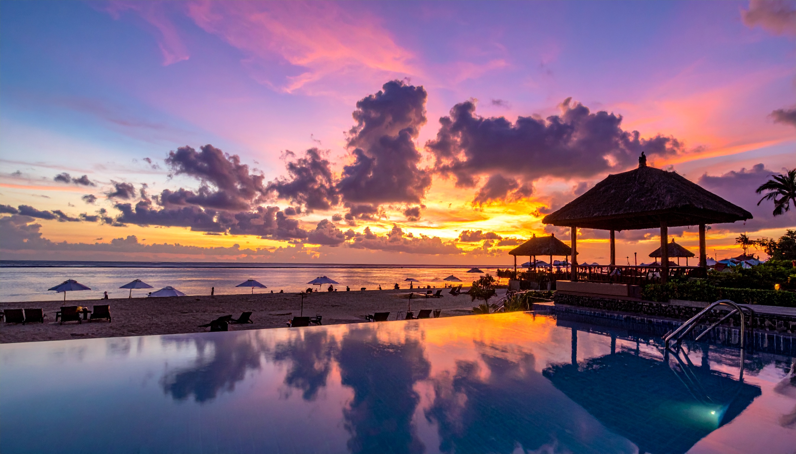 Pôr do sol deslumbrante em uma praia tropical com piscina de borda infinita. Ideal para promover destinos de férias e resorts de luxo, a imagem captura um ambiente relaxante e sofisticado. A cena é dominada por tons vibrantes de roxo e laranja, refletidos tanto na água quanto nas nuvens. Perfeito para uso em campanhas de marketing de turismo e hospitalidade durante a temporada de verão. A combinação de elementos naturais e estruturas de lazer destaca o equilíbrio entre conforto e beleza natural.