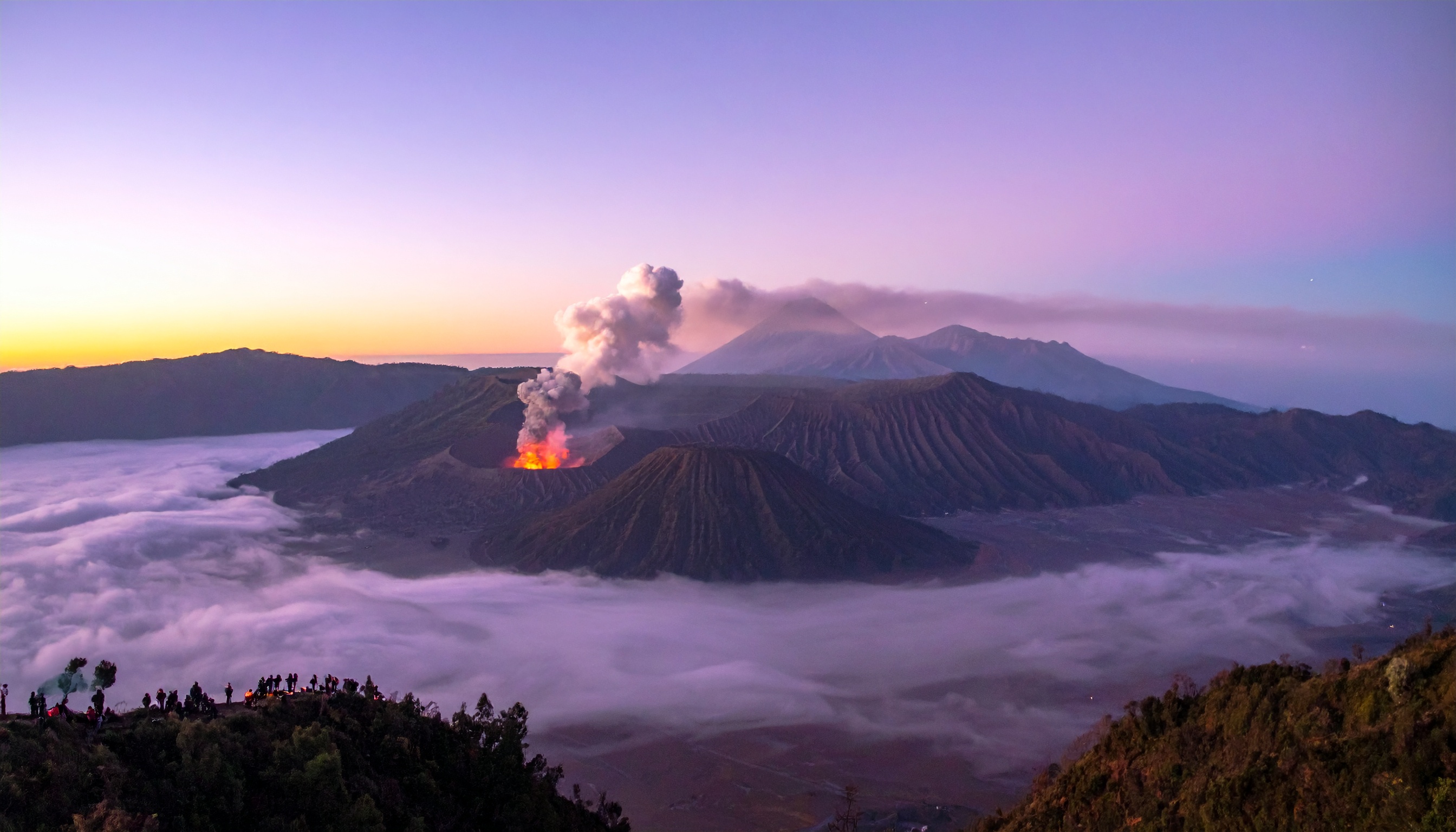 O Monte Bromo exibe sua majestosa erupção durante o amanhecer, cercado por um mar de nuvens. Este cenário espetacular é perfeito para amantes da natureza e fotografia de paisagens. As cores vibrantes do céu e o brilho da lava criam um contraste impressionante. Ideal para uso em materiais promocionais de turismo e aventura. A imagem captura a beleza e a força da natureza, evocando um senso de admiração e respeito.