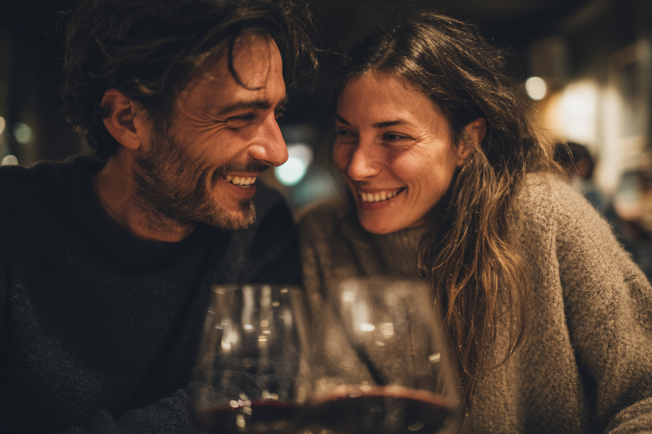 Casal sorrindo e se divertindo em um jantar romântico, com taças de vinho tinto à frente em um ambiente aconchegante e iluminado.