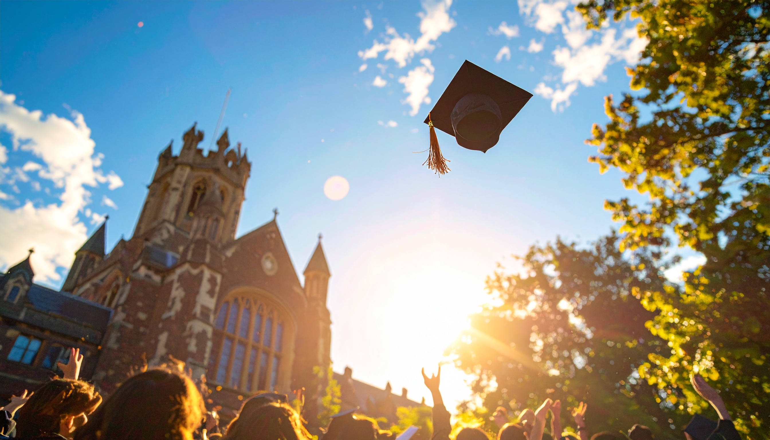 Cena vibrante de formatura ao ar livre, com capelo lançado ao ar contra um céu azul e ensolarado. A arquitetura gótica de uma universidade ao fundo, cercada por árvores verdes, destaca-se na iluminação dourada do pôr do sol. A multidão, em silhueta, celebra o momento, criando uma atmosfera de alegria e conquista.