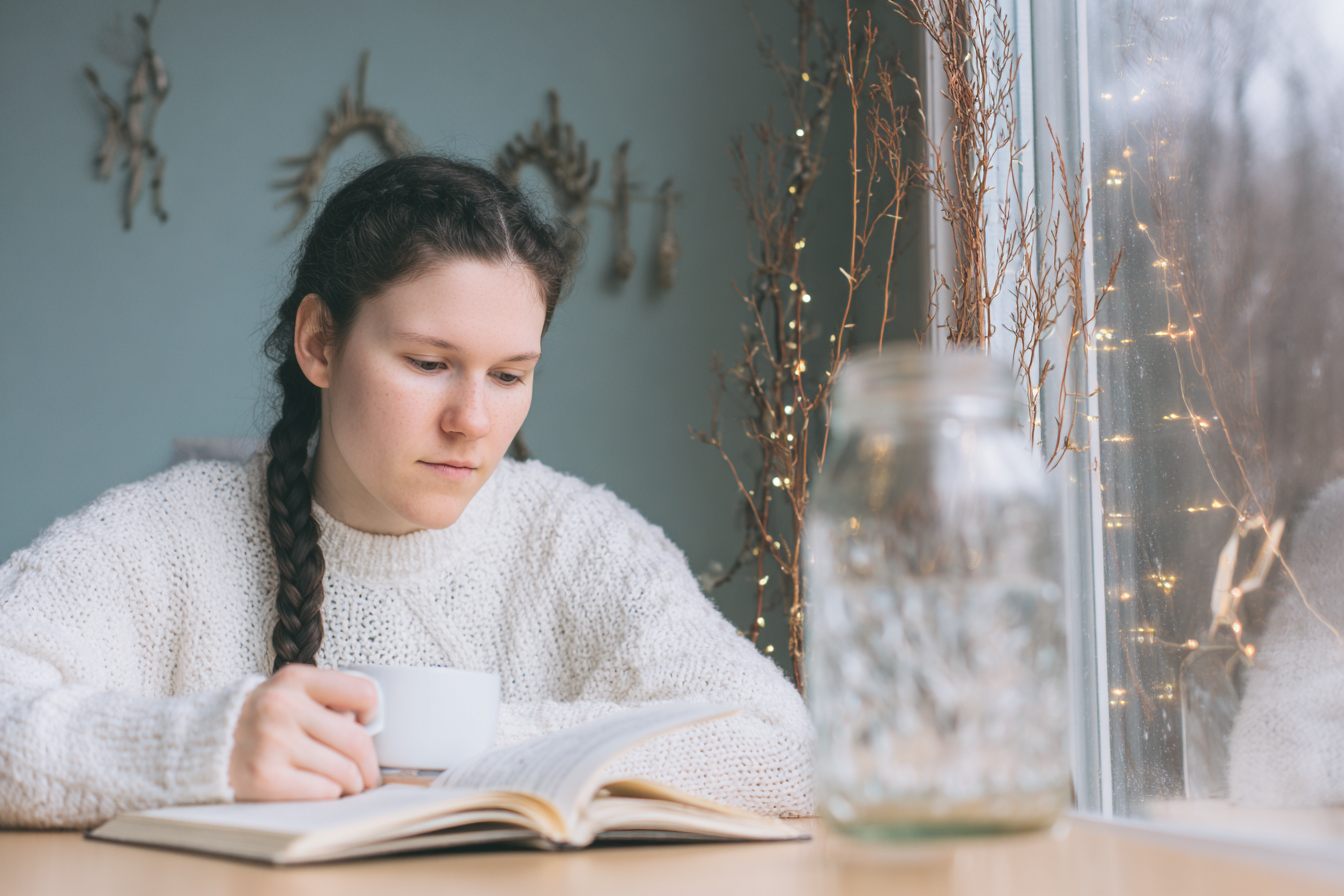 Jovem mulher com tranças longas em suéter branco, segurando xícara de café enquanto lê livro em mesa de madeira. Luz suave entra pela janela decorada com galhos e luzes piscantes, criando atmosfera de tranquilidade e introspecção. Composição equilibrada com foco difuso ao fundo.