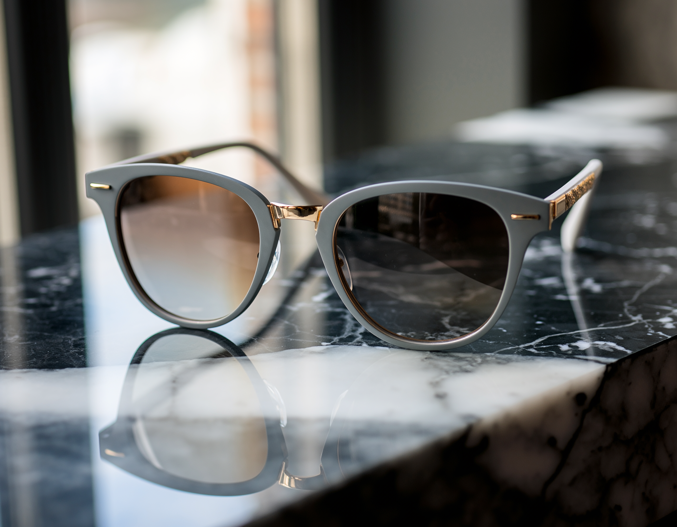 Óculos de sol com armação cinza claro e detalhes dourados sobre superfície de mármore preto com veios brancos, refletindo suavemente a luz natural ambiente.