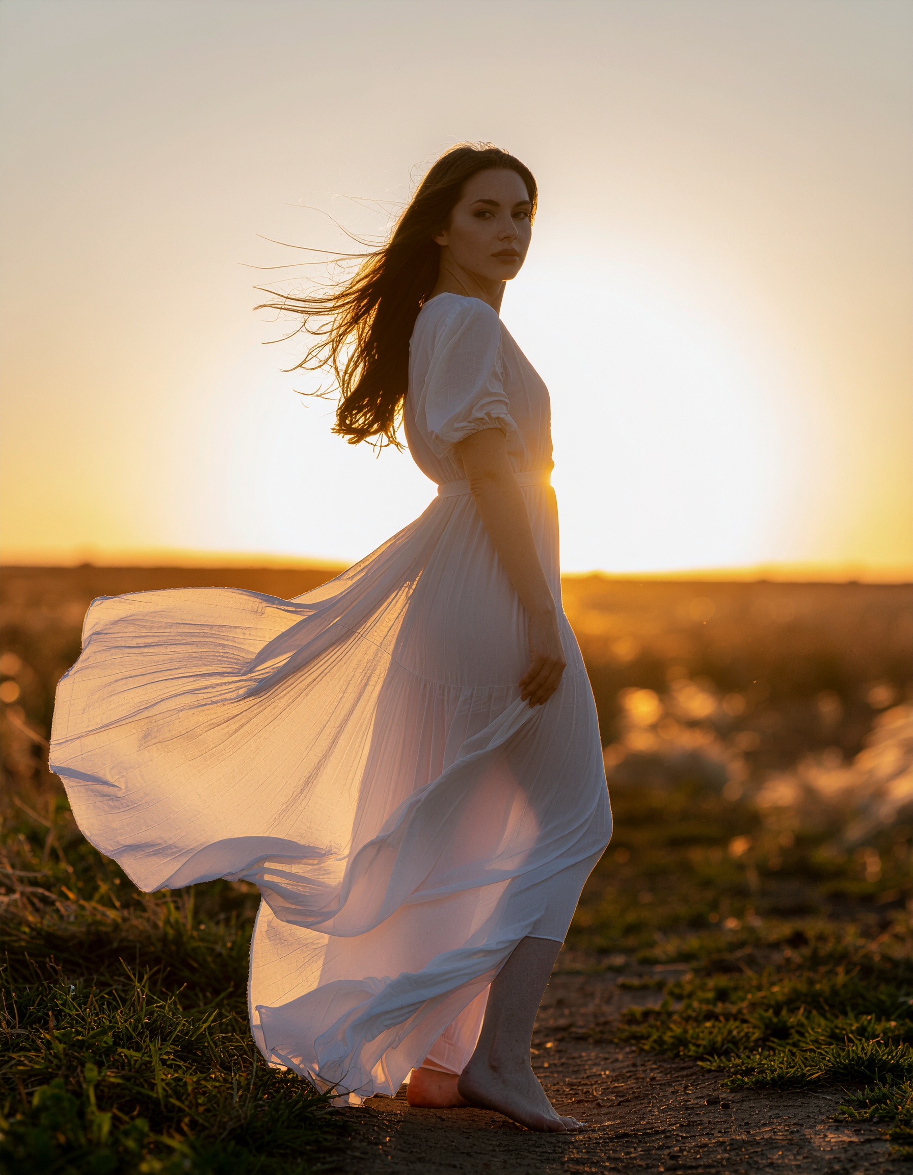 A woman stands in a flowing white dress, backlit by a golden sunset
