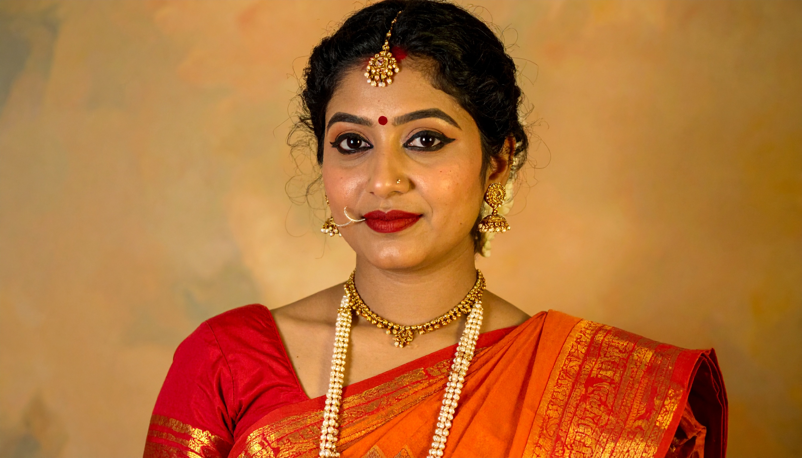 A woman elegantly dressed in traditional Indian attire with intricate jewelry stands against a warm