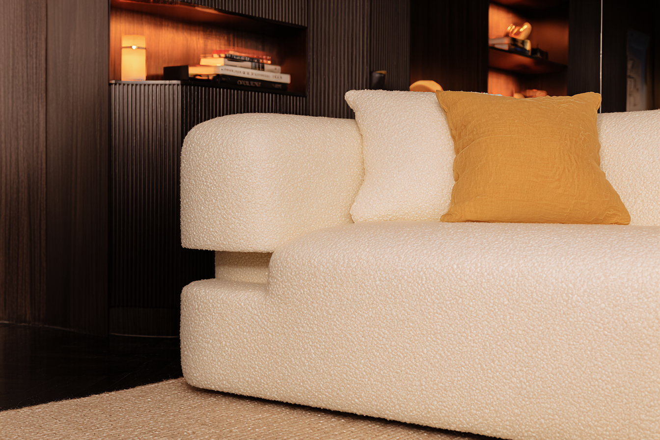 Um sofá moderno com textura de bouclé em tom creme, complementado por uma almofada amarela mostarda, em uma sala de estar elegantemente iluminada com prateleiras embutidas e livros decorativos, transmitindo uma sensação de aconchego e sofisticação minimalista.