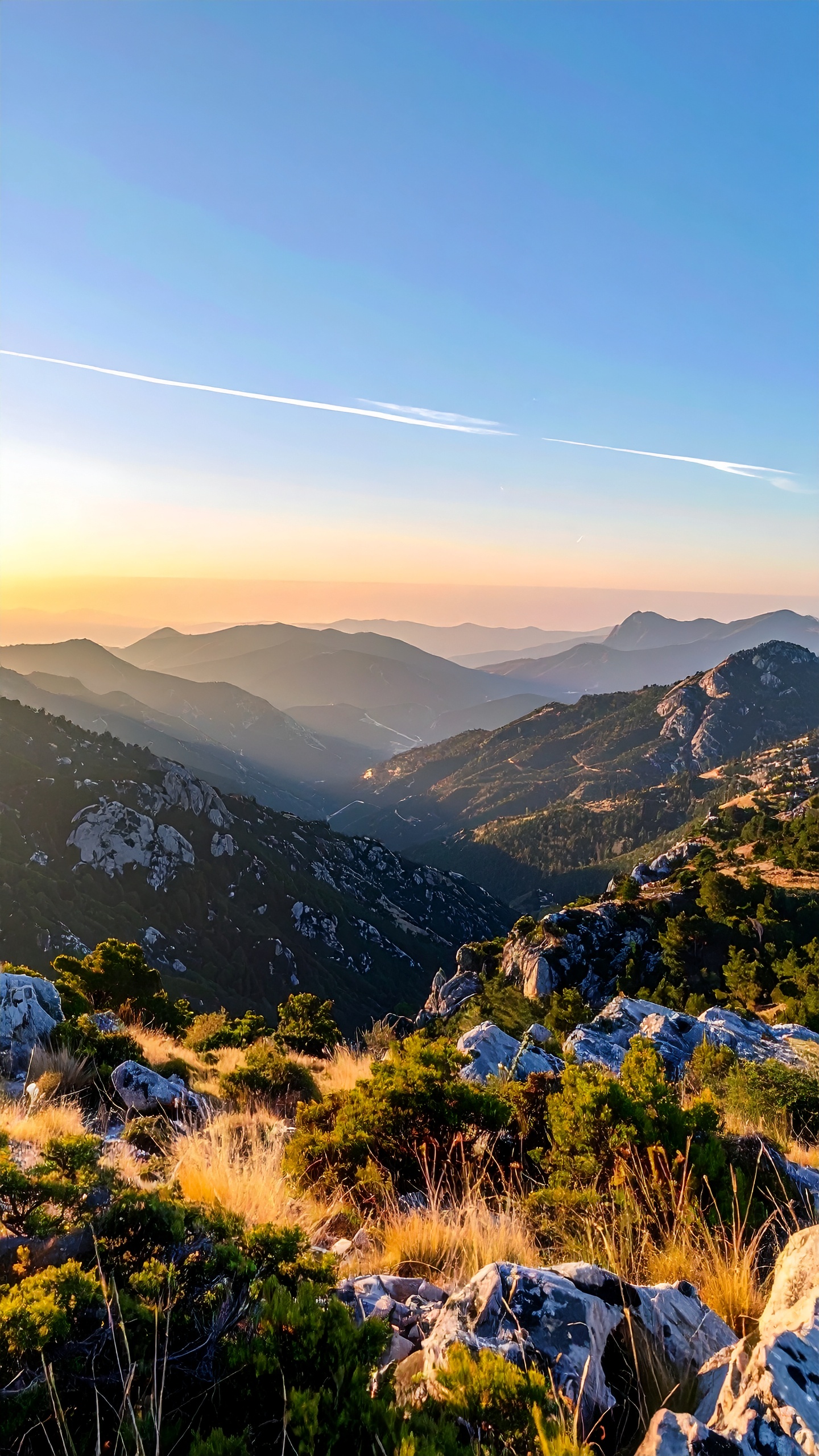Paisagem montanhosa ao pôr do sol, com camadas de montanhas azuladas se estendendo até o horizonte. Vegetação rasteira e rochas em primeiro plano iluminadas por luz dourada. Céu limpo com tons de azul e laranja, criando uma atmosfera serena e majestosa. Perspectiva ampla e profunda, capturando a vastidão e beleza natural do cenário.