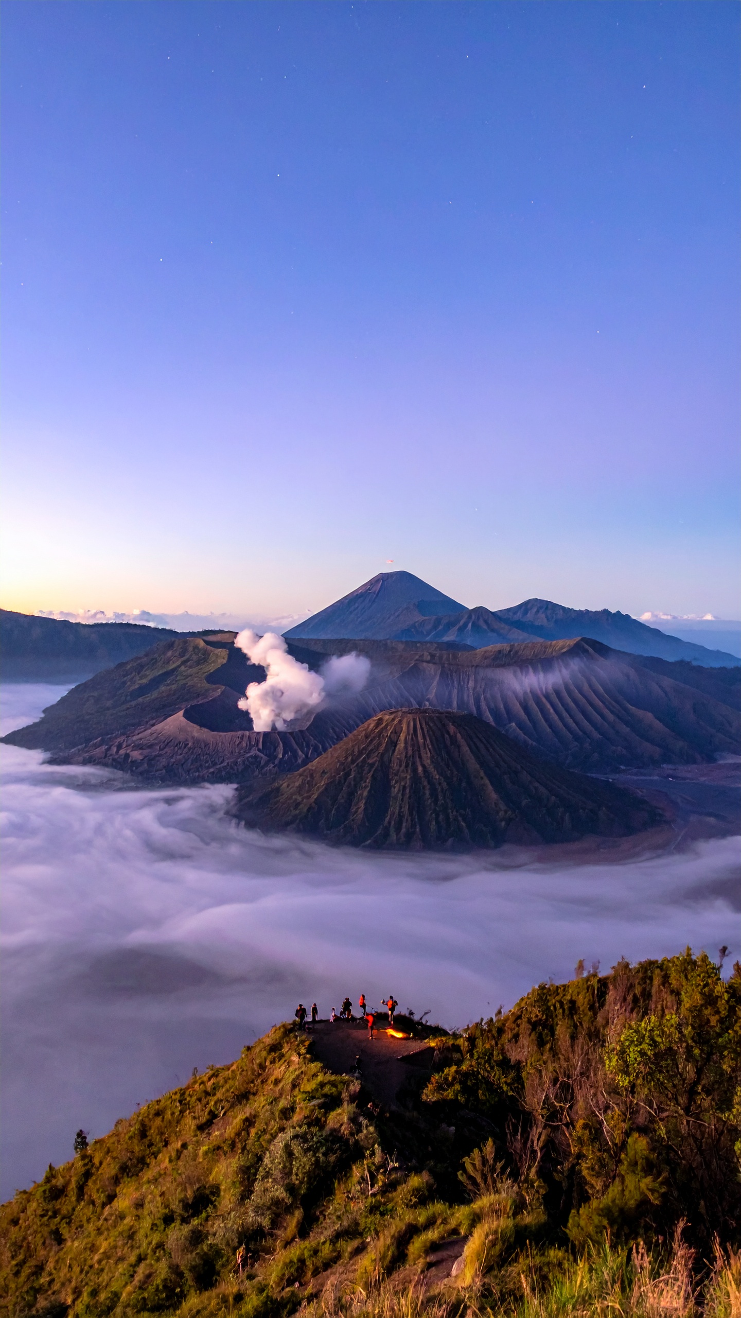 Vulcão Bromo cercado por nuvens ao amanhecer em Java, Indonésia. Este local é ideal para trilhas e aventuras fotográficas, oferecendo vistas inesquecíveis. O contraste entre o céu azul suave e o vapor do vulcão cria uma cena espetacular. Visitar de manhã cedo proporciona uma experiência única com a natureza. A região é um ponto turístico popular, conhecido pela sua beleza natural impactante.