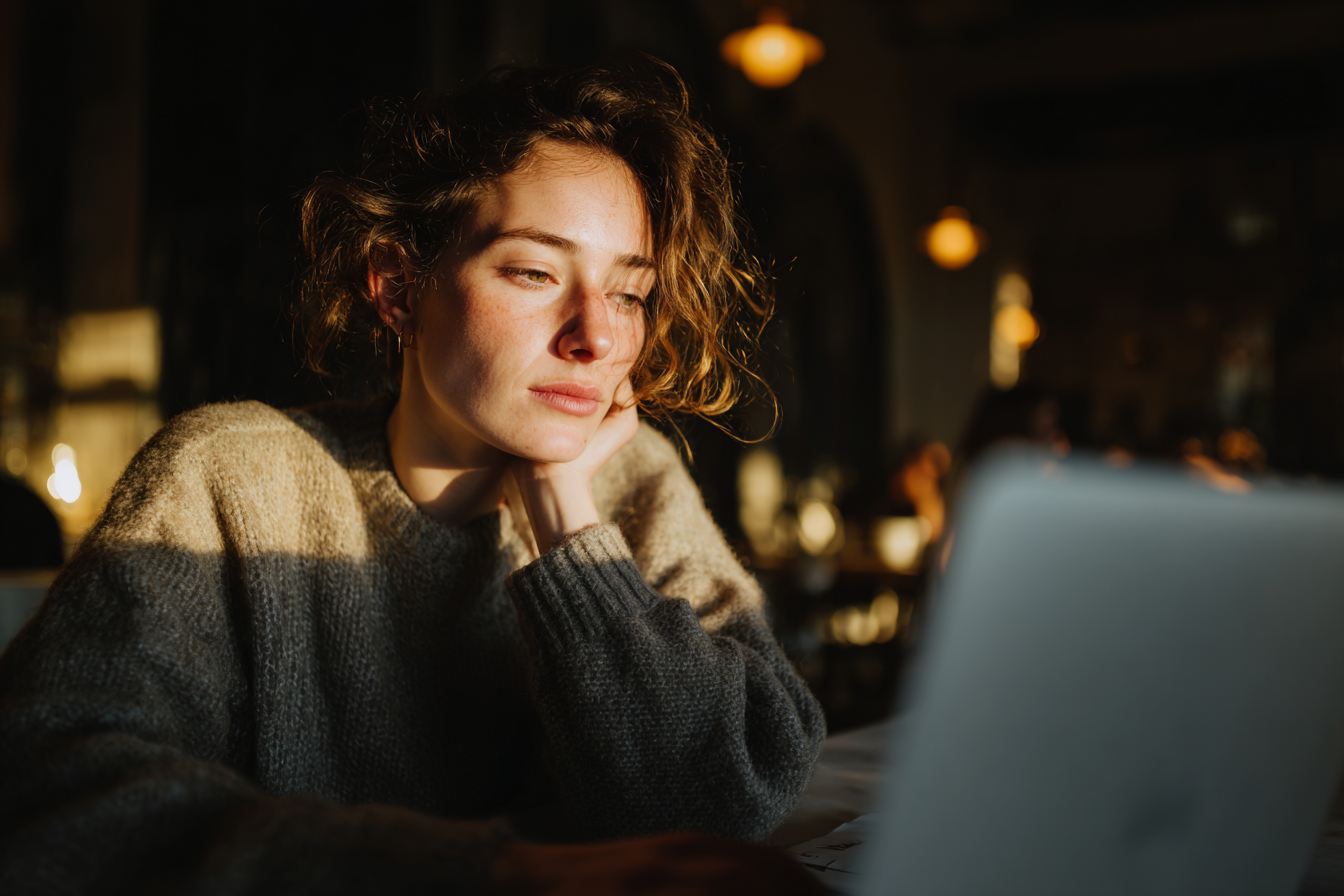Jovem mulher pensativa em um café iluminado pelo sol, trabalhando em um laptop. Atmosfera acolhedora e reflexiva com luz suave e tons quentes.